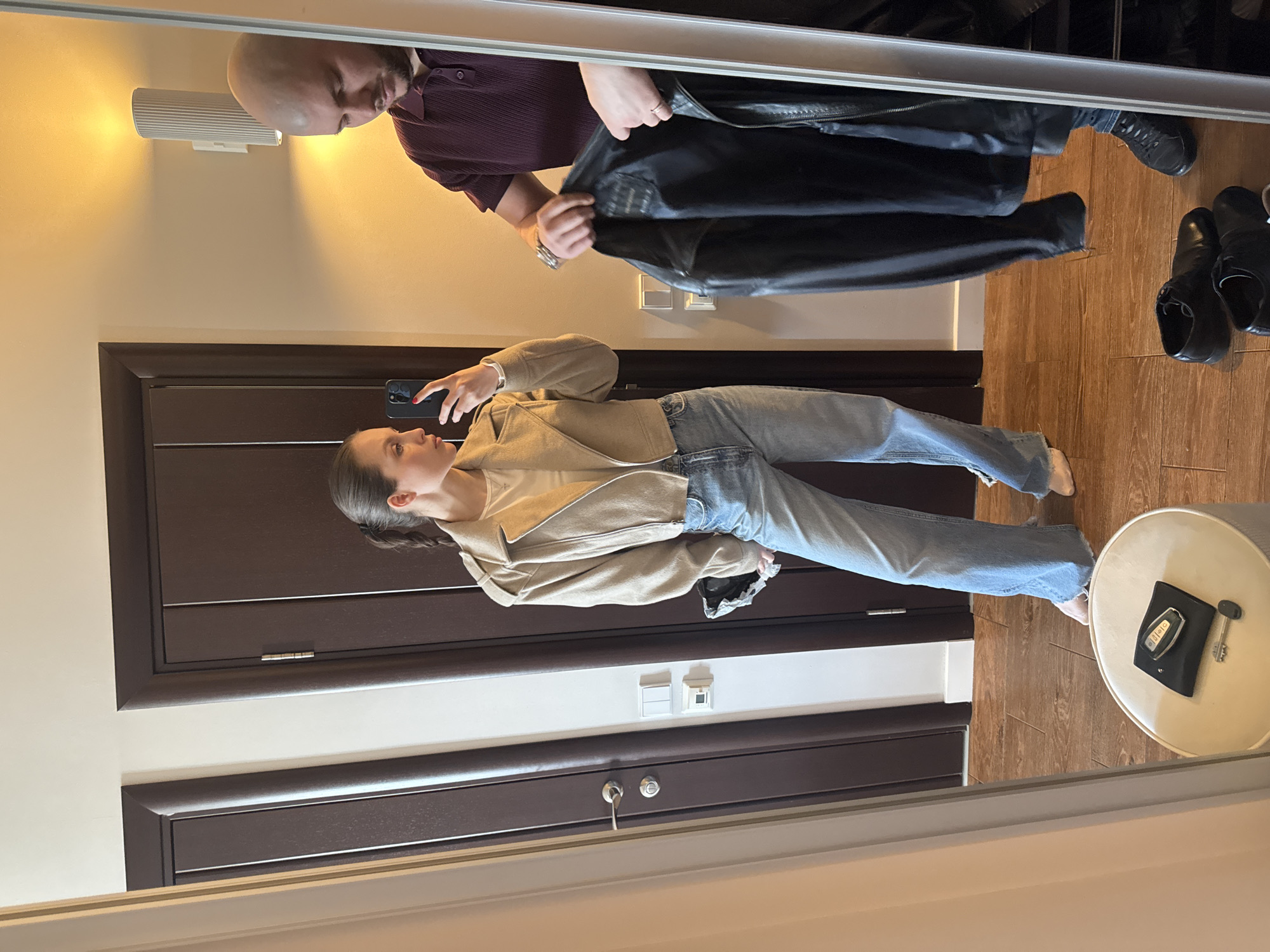 A mirror image shows a young woman in a light brown jacket and blue jeans taking a picture with her cell phone. A bald man in a maroon shirt holds a black jacket behind her. Shoes are on the floor near a small table. The setting seems to be an entryway in a home. It's impossible to pinpoint the exact location in Moscow or Russia from the photo alone.