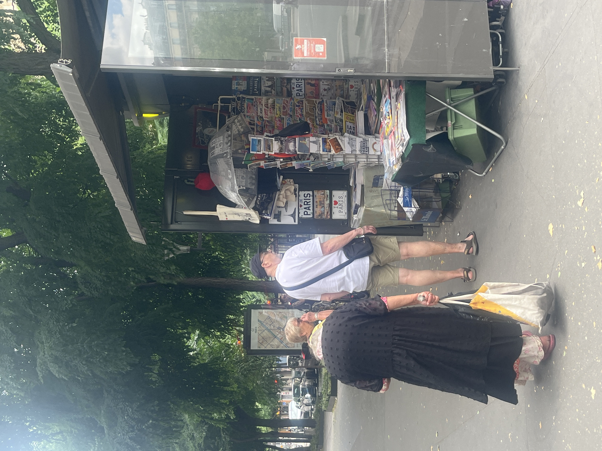 In Paris, France, an outdoor kiosk selling newspapers and souvenirs is displayed. A man and two women are in front of the kiosk. The man is wearing a white t-shirt, khaki shorts, and a black cap. He is looking to the right. One woman is wearing a black dress and a white and yellow tote bag. The other woman is wearing a yellow shirt and a pink scarf. She is looking at the man. The kiosk has an assortment of items, including postcards, magazines, and an umbrella. There are trees in the background and cars on the street.