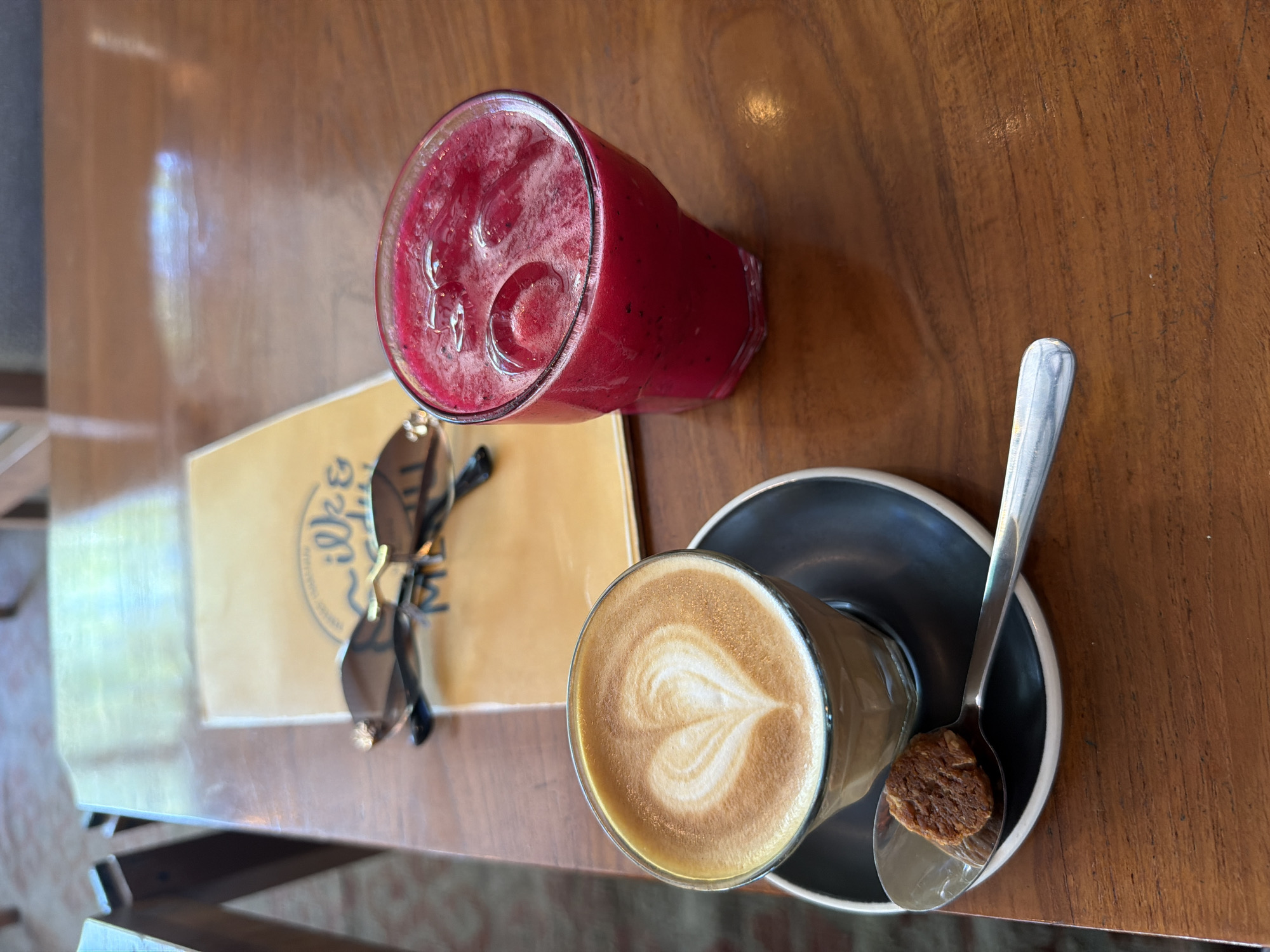 The image depicts a table setting with a red beverage, a latte with a heart design, sunglasses, and a menu card. The red beverage is in a clear glass with ice and appears to be a smoothie or juice. The latte is in a similar clear glass and has been topped with steamed milk in a heart shape, resting on a black saucer with a silver spoon and a small cookie. The sunglasses are dark-rimmed and are placed on top of a yellow menu card with a logo or text, potentially indicating the cafe's name. The table is wooden with a warm tone and visible grain. The scene is situated in a cafe or restaurant, and based on the illy logo on the menu card, it is most likely located in the city of Unknown, Unknown. The time of day is likely daytime.