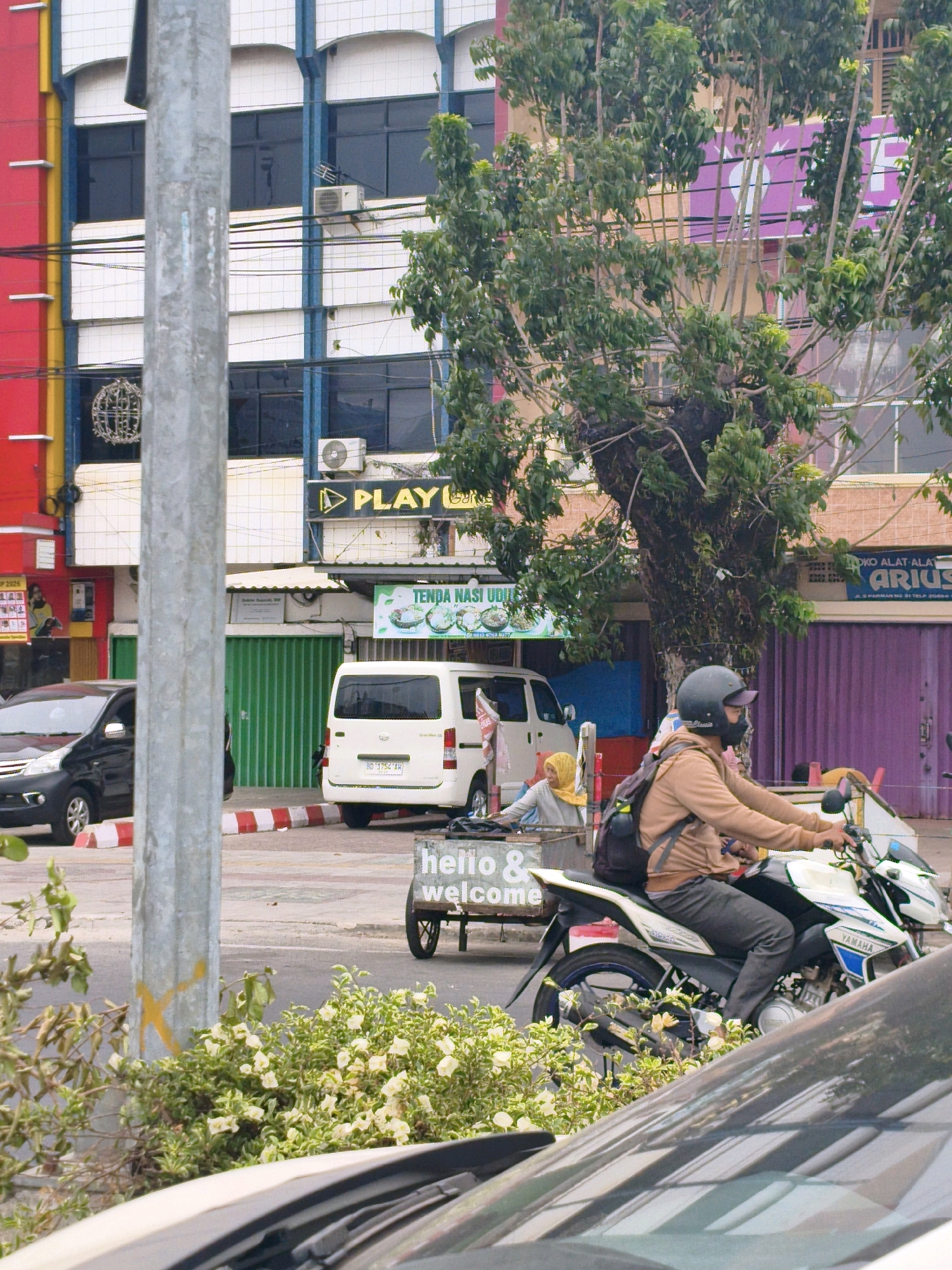 In the city of Bengkulu, Indonesia, a person wearing a helmet and a tan jacket is riding a white and blue Yamaha motorcycle. The motorcycle is towing a small trailer with the words "hello & welcome" written on it in white text. Behind the motorcycle, a white van is parked, and next to it is a food stall with a green awning. The scene is set against a backdrop of multi-story buildings with brightly colored facades, including red, yellow, and blue. A large tree with dense green foliage partially obscures the buildings. The street is paved, and there are parked cars visible on the left side of the frame. The overall impression is a bustling street scene in an urban environment.