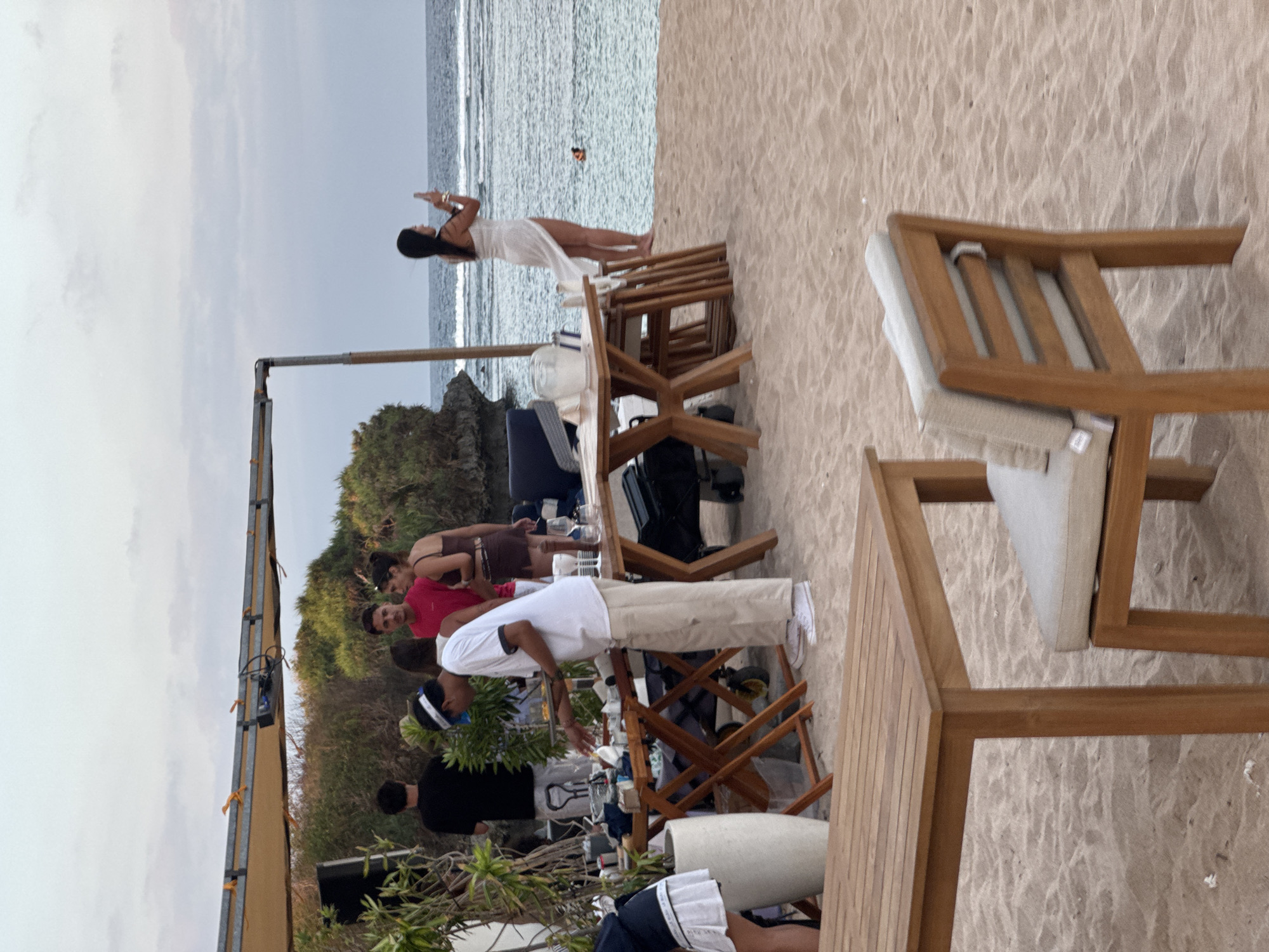 The image depicts a beach scene in Kuta Selatan, Indonesia, with a group of people gathered near a setup of tables and chairs on the sand. In the foreground, two wooden tables with striped surfaces are visible, one with a chair featuring a beige cushion. Behind these, a cluster of wooden tables with chairs are arranged, some with white cloth coverings.
Several individuals are interacting around these tables. A person in a white shirt and beige pants is tending to food laid out on a table. To their right, a woman in a pink top and a woman in a brown bikini are engaged in conversation. Further back, a man in a black shirt stands with his back to the camera.
In the middle ground, a woman in a white dress stands near the edge of the beach, holding a phone and seemingly taking a picture of the ocean. The ocean is calm with gentle waves, and a small orange buoy is visible in the water. The background features lush green vegetation on a rocky outcrop and a cloudy sky. A large, light-colored tent or canopy is stretched over the gathering area, supported by metal poles. The overall atmosphere suggests an outdoor dining or event setup on a tropical beach.