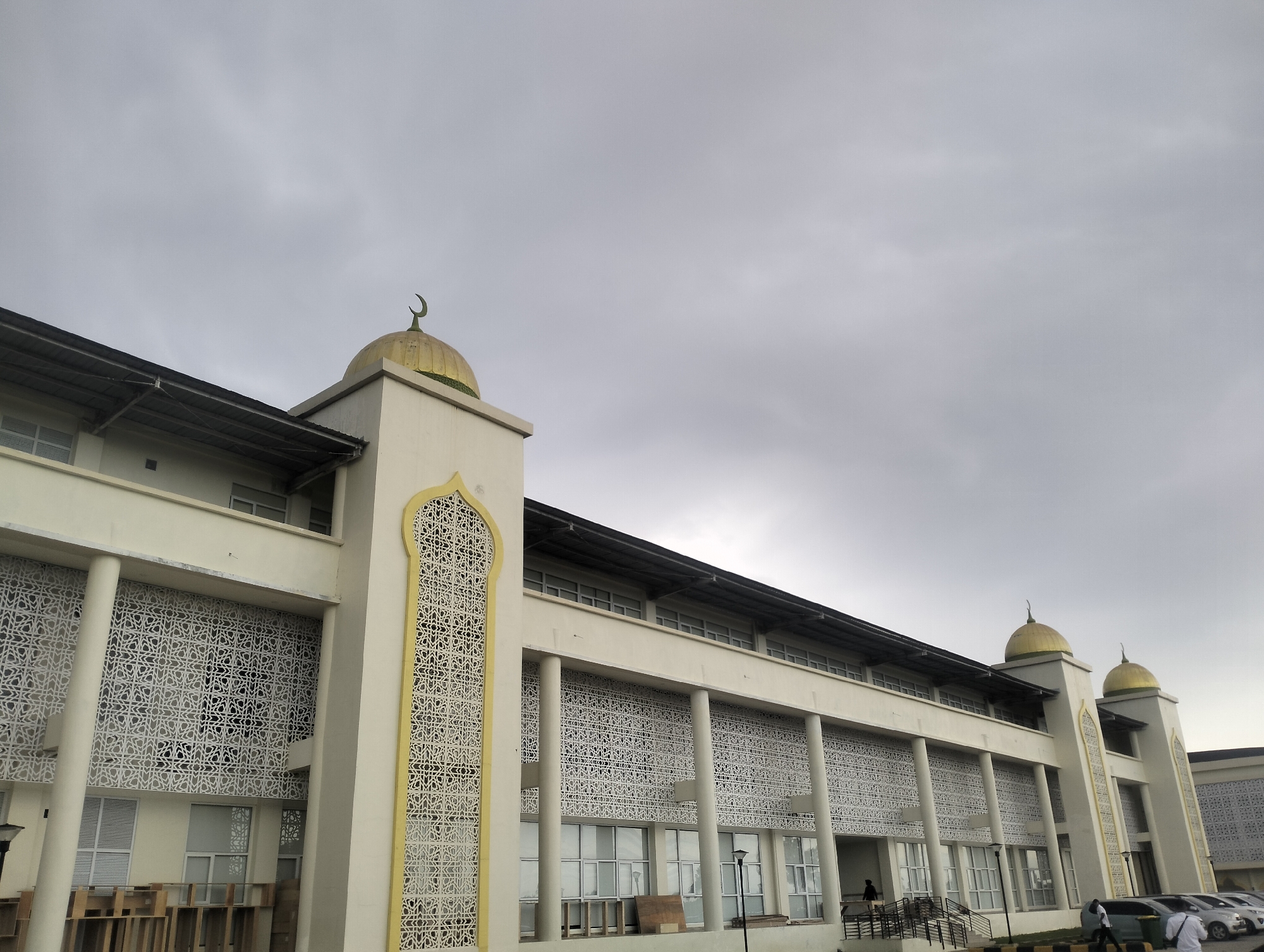 The image shows the exterior of a large, modern building with Islamic architectural elements, specifically a mosque, in Reuleut Timu, Indonesia. The building is primarily cream-colored with ornate white screens featuring Islamic geometric patterns on its facade. Two golden domes, each topped with a crescent moon, are visible on the roof. A long row of windows runs across the building's upper level, supported by white columns. The sky is overcast, and the lighting suggests it is either early morning or late afternoon. In the foreground, several parked cars are visible, and a few people are present, including one person walking near the entrance stairs and another near the parked cars. Wooden structures are stacked in front of some windows on the lower level. The overall impression is of a significant public building set against a muted sky.