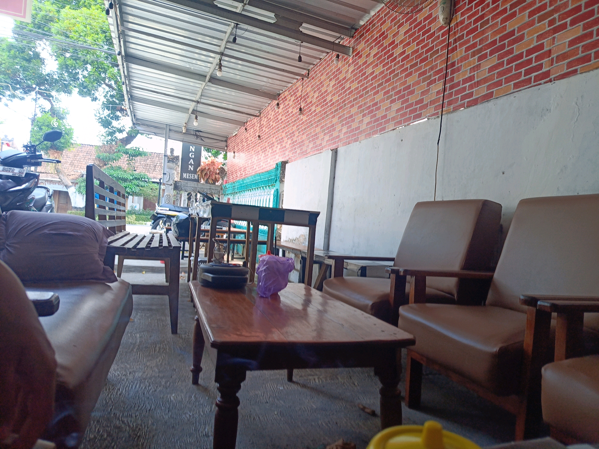 The image depicts an outdoor seating area, likely a cafe or warung, in Candirejo, Indonesia. The area is covered by a corrugated metal roof supported by metal beams, with exposed light bulbs hanging from it. The back wall is adorned with faux brick tiling in shades of red and orange, transitioning to a plain white section lower down.
In the foreground, a low wooden coffee table with turned legs holds a black circular object and a crumpled purple plastic bag. To the right, two brown leather armchairs with wooden frames are positioned. To the left and further back, a long wooden bench with a slatted seat is visible, with a dark-colored pillow placed on it. Behind the bench, a motorcycle is parked.
In the mid-ground, other seating arrangements, including wooden chairs and tables, are present. A sign with vertical text, likely in Indonesian, is mounted on a pole, and a potted plant with reddish-orange foliage sits nearby. The ground is paved with textured concrete. In the background, trees and buildings with tiled roofs suggest a residential or semi-commercial area. The overall impression is of a casual, open-air gathering spot.