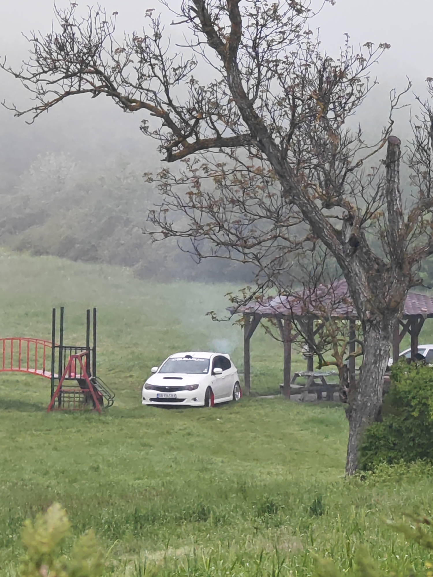 The image depicts a grassy area, likely a park or open space, under an overcast sky, possibly in Shumen, Bulgaria, based on the license plate of the white Subaru Impreza WRX STi parked on the grass. A red and grey children's playground slide is visible to the left of the car. In the background, there is a wooden gazebo with a picnic table underneath. The atmospheric conditions suggest mist or fog. The trees in the foreground are leafless and have distinctive branching patterns. Smoke is visible coming from the car's exhaust pipe. Another car is faintly visible parked behind the gazebo. The overall scene is tranquil and possibly rural.