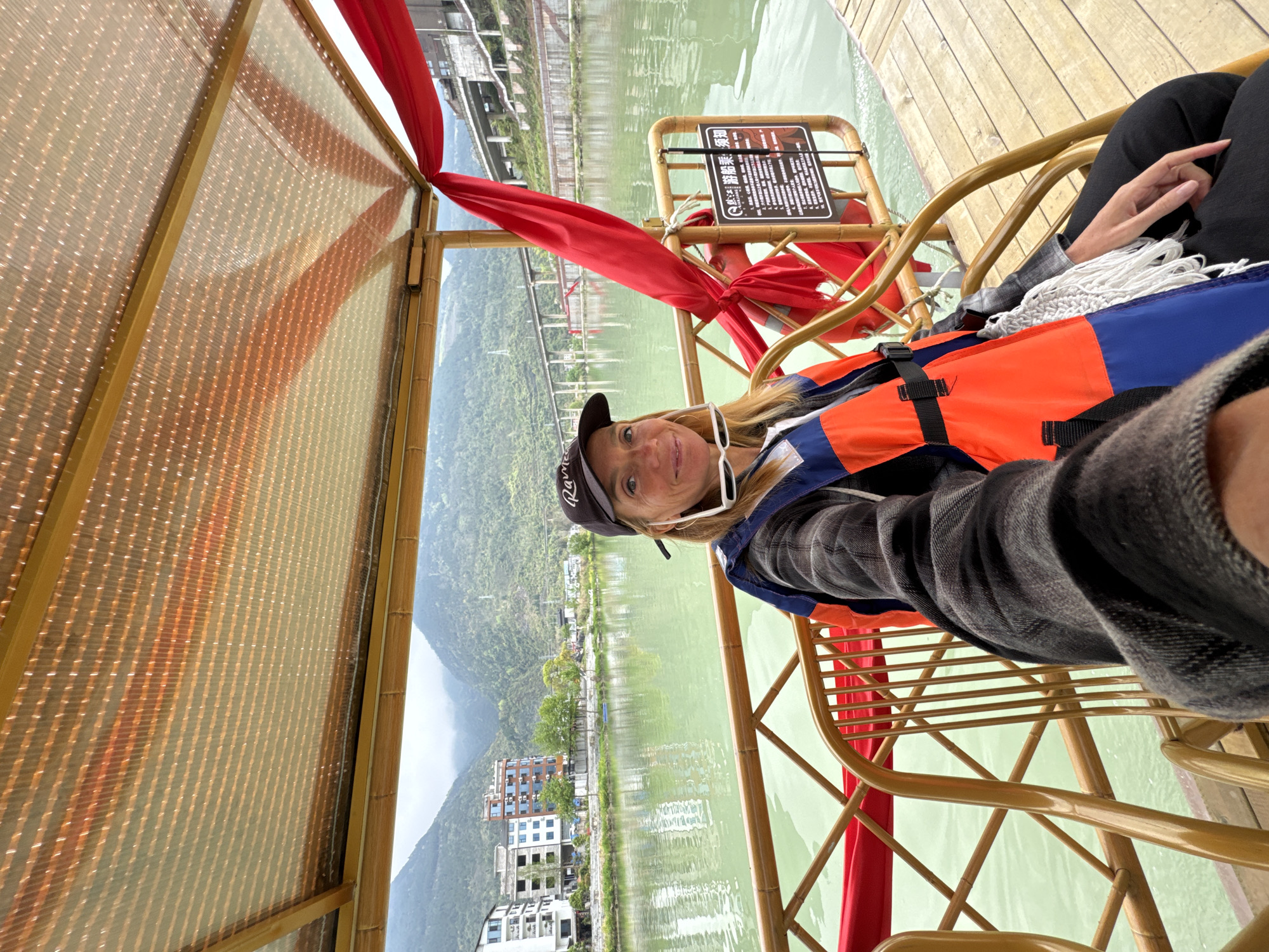 A woman wears a life vest and a baseball cap while seated in a boat on a body of water. The boat has a canopy above and a red decorative fabric. Buildings and a hillside can be seen along the water's edge. The location appears to be in the city of Zhuoshui, China.