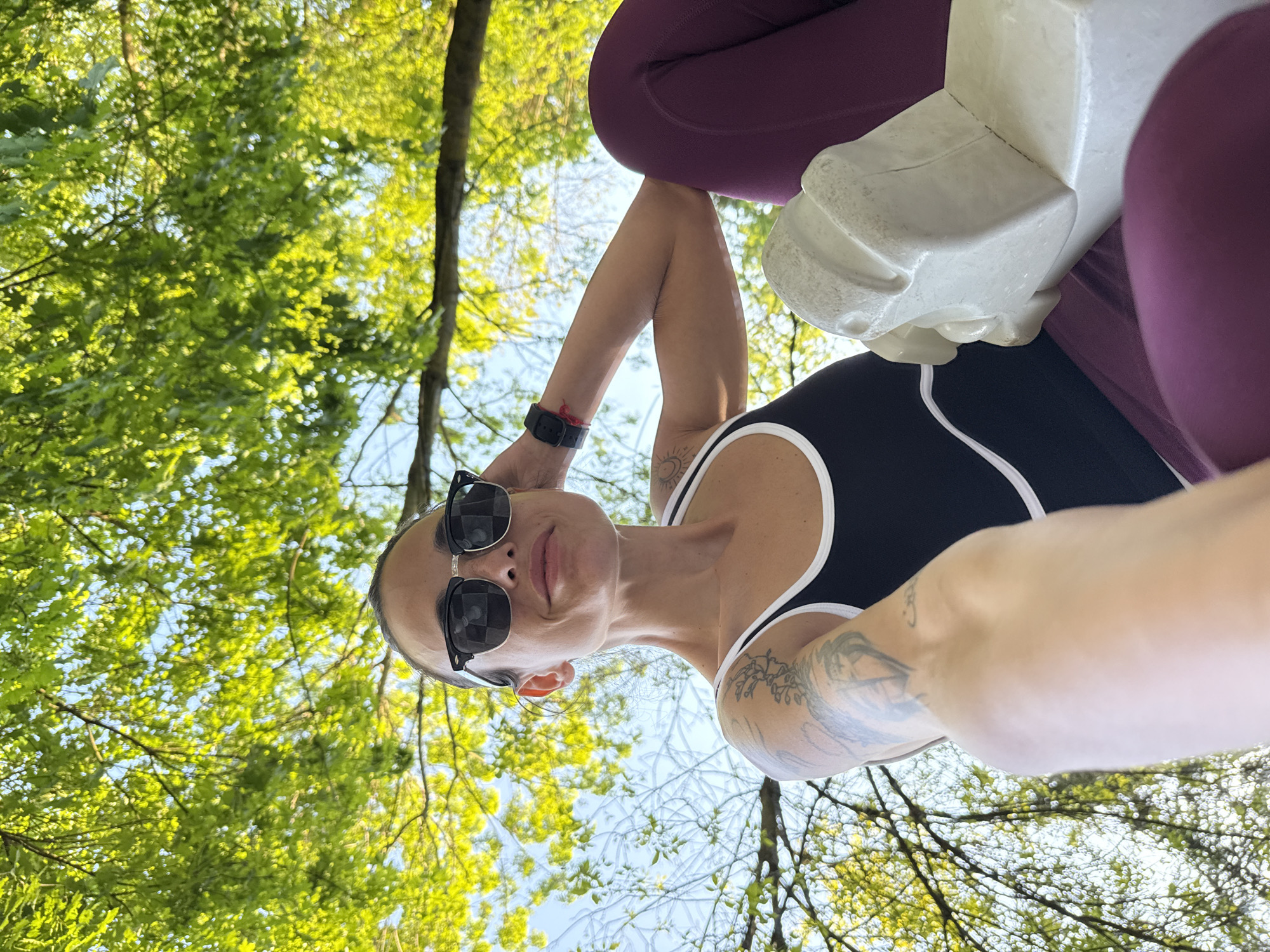 A low-angle shot looking up at a woman in a black and white tank top and burgundy leggings. She is holding what appears to be a white structure or object, possibly made of plaster or a similar material. She is wearing sunglasses, and there are visible tattoos on her arm and chest. The background consists of trees with green leaves and a patch of blue sky visible through the foliage. The location is Pleven, Bulgaria.