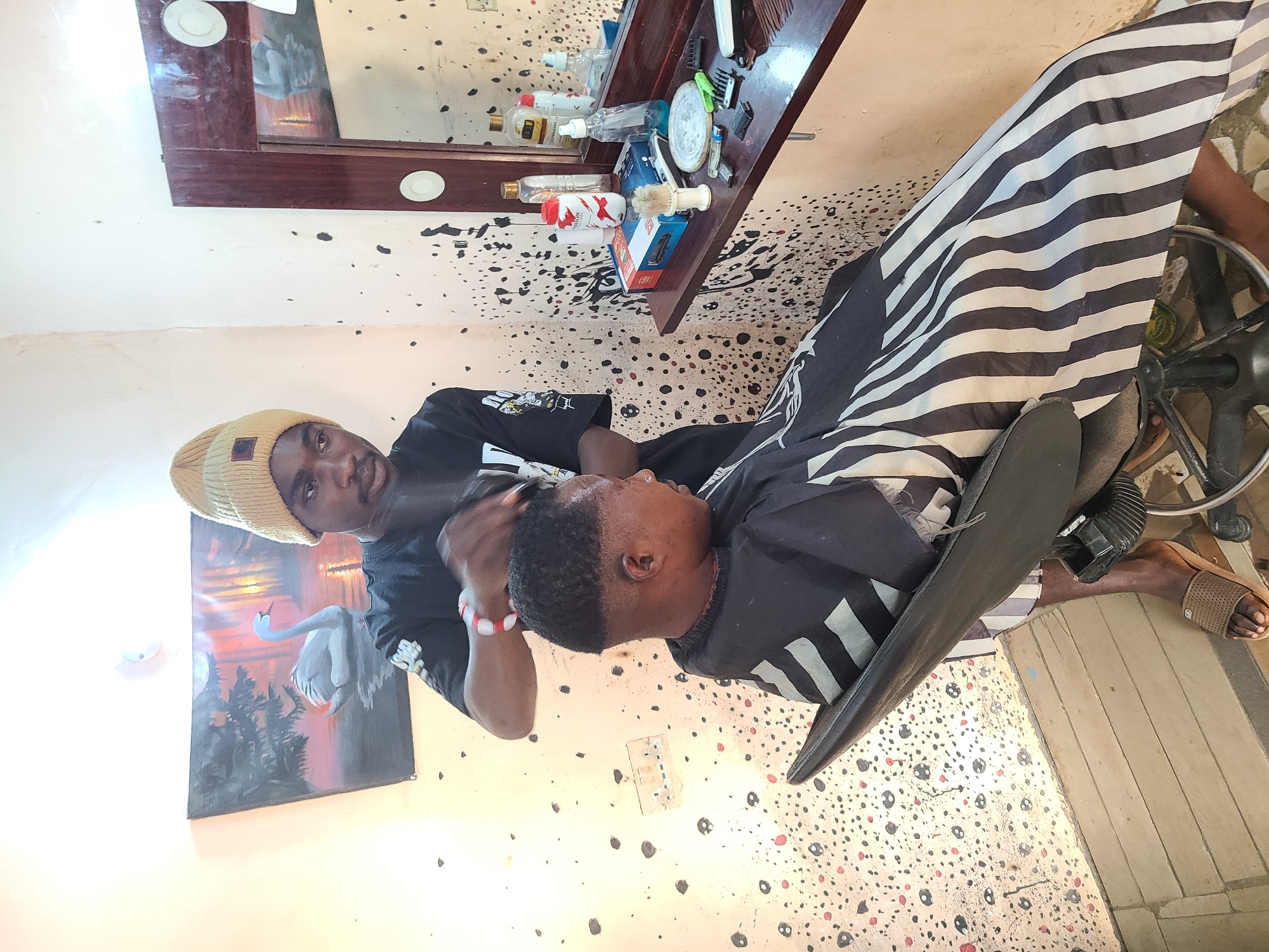 A barber is cutting a customer's hair. The barber, wearing a tan beanie and a black shirt, stands behind the seated customer. The customer is draped in a black and white striped barber's cape. The environment suggests a barber shop, with a mirror and a shelf of barbering supplies visible in the background. The floor is patterned with splatters. The location is plausibly Jos, Nigeria, given the attire and physical appearance of the individuals.