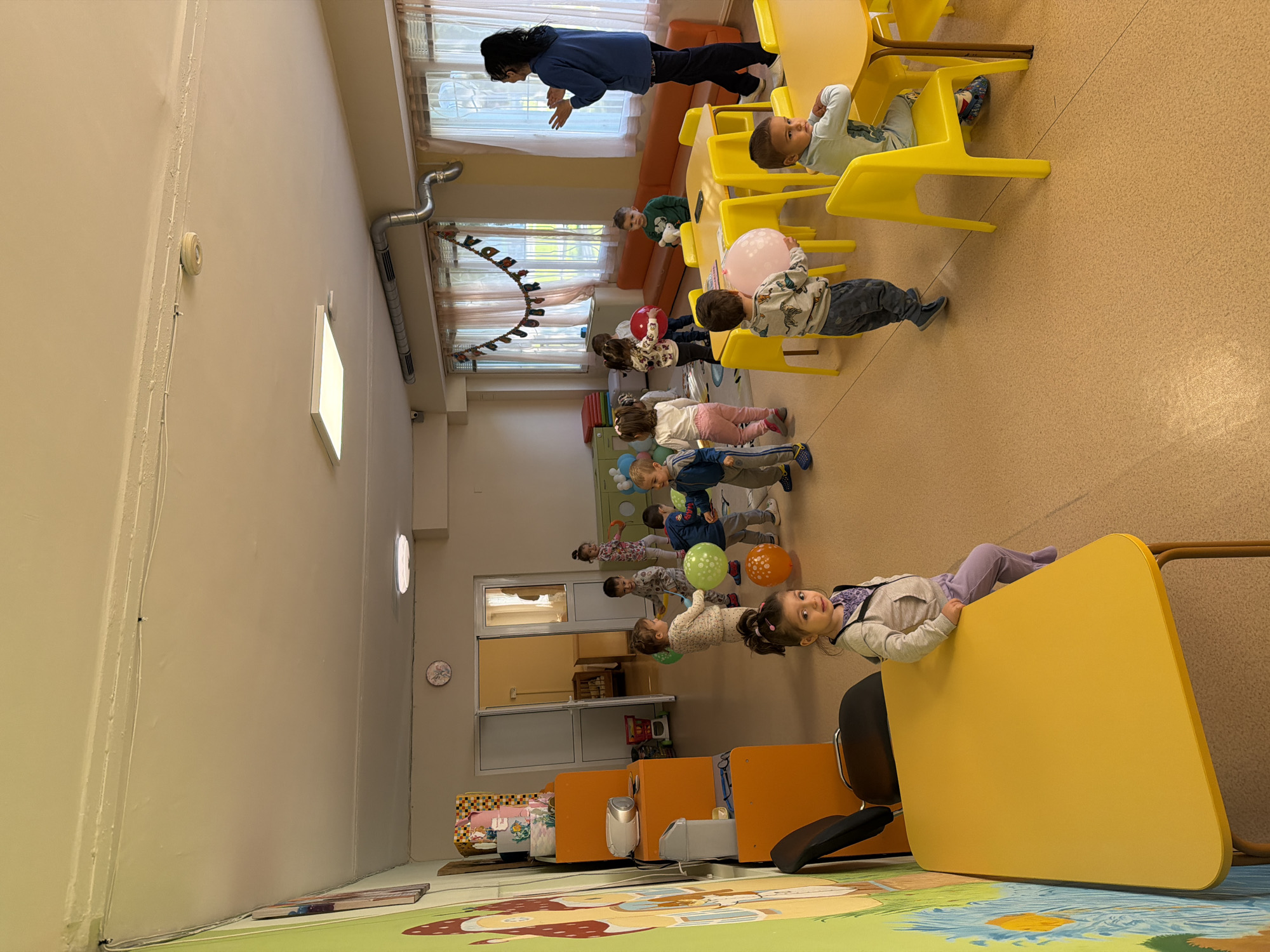 The image depicts a group of young children in what appears to be a classroom or daycare setting, likely in Burgas, Bulgaria. The children are arranged in a line, some holding balloons. An adult is interacting with the children. The room has yellow chairs and tables, a colorful mural on the lower part of the wall, and a decorative garland. A girl is reclining on a yellow table, looking at the camera. The setting is indoors.