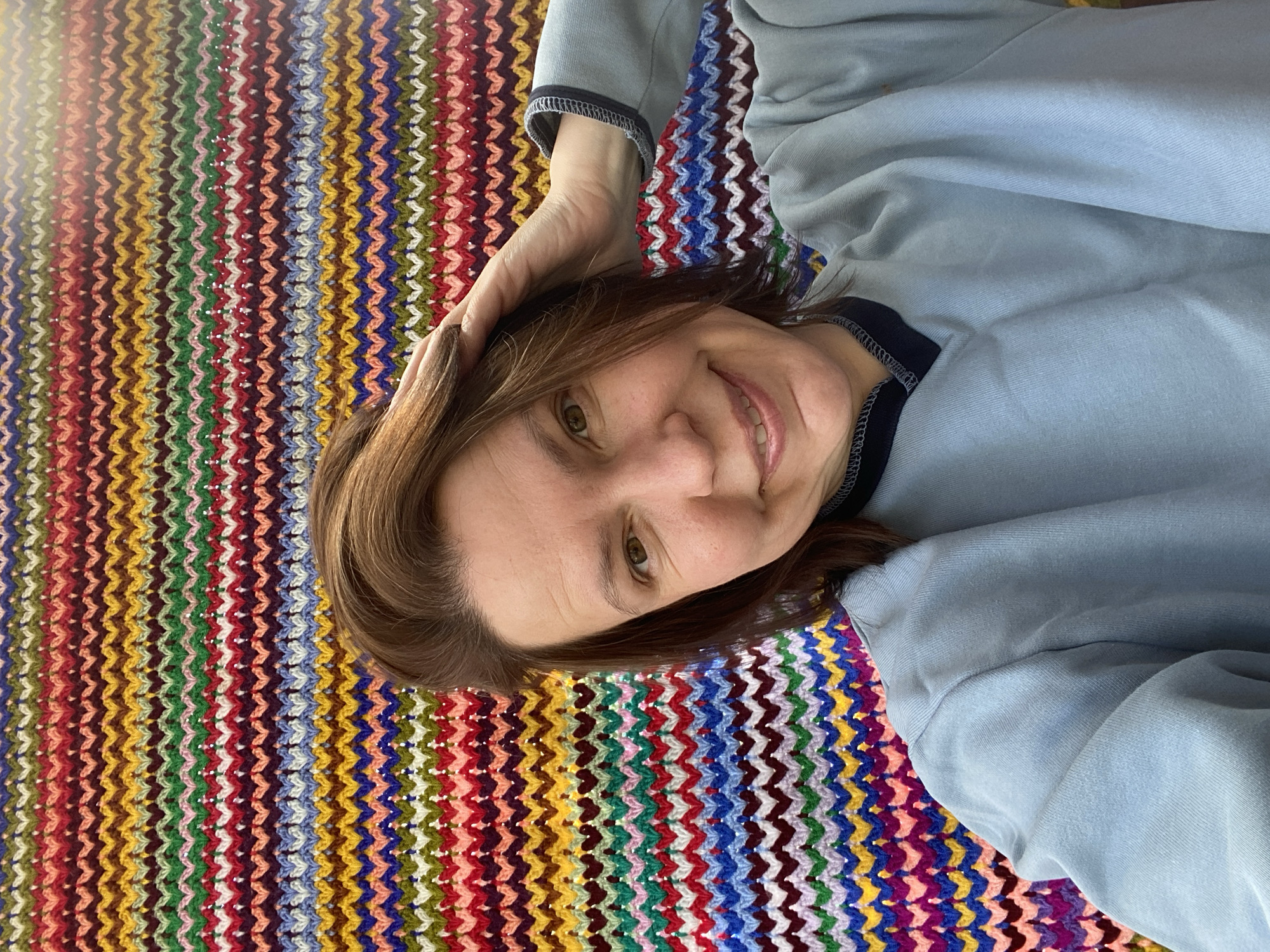 A woman with brown hair lies on a colorful zig-zag patterned surface. She wears a light blue top with dark blue trim. Her left hand rests on her forehead. She smiles slightly and looks at the camera. The pattern beneath her has stripes of red, orange, yellow, green, blue, and purple in a chevron design.