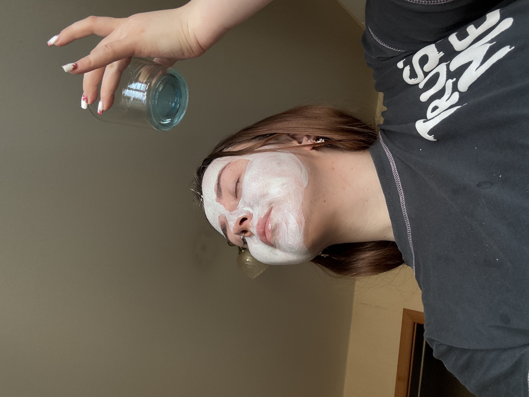 A woman with a white face mask is lying down while someone holds a glass jar above her face. The woman has light brown hair. The person holding the jar has painted nails. The woman is wearing a black shirt with white lettering. The location is not recognizable as Izhevsk, Russia. The background is a beige wall with a door.