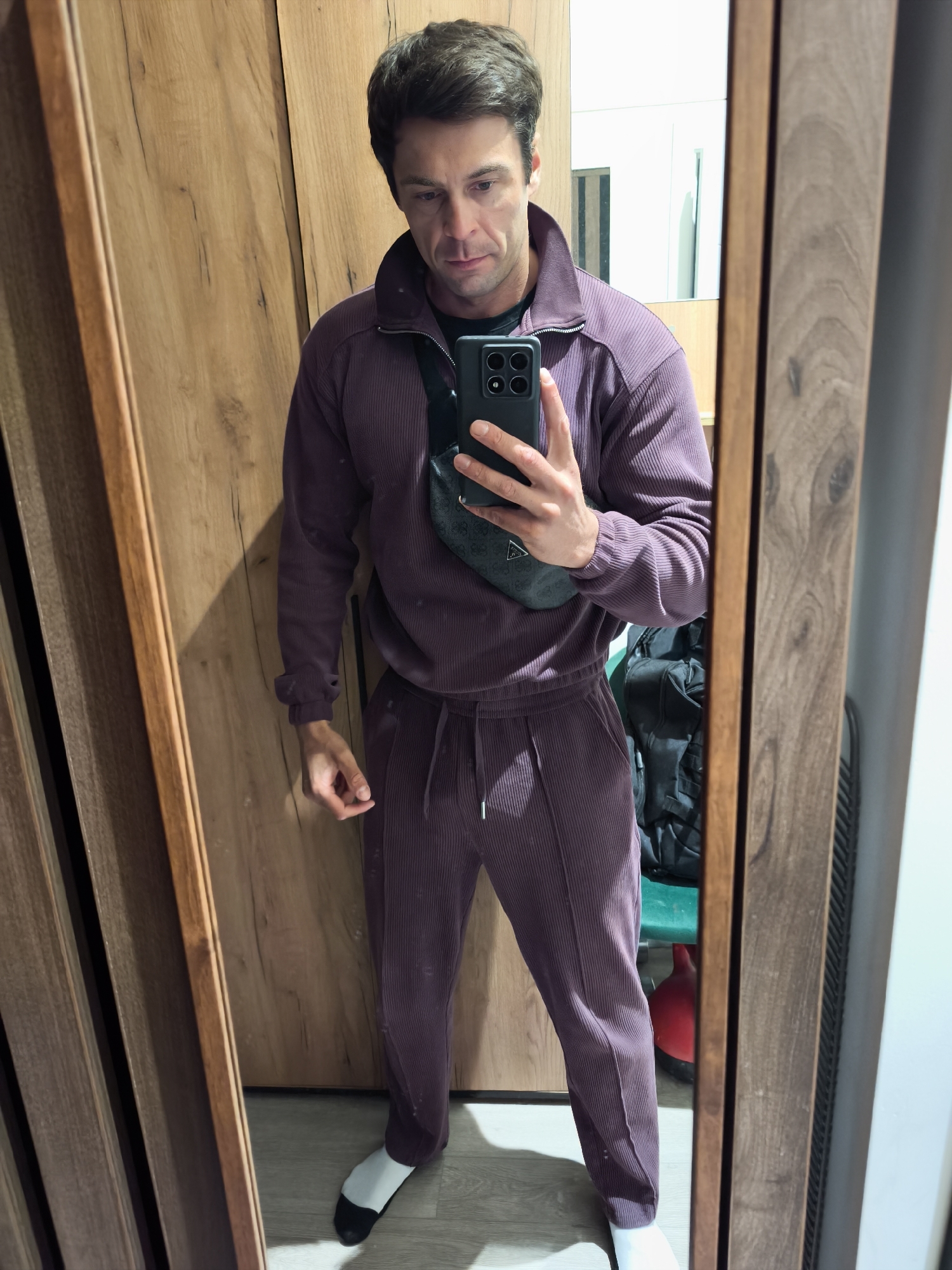 A man stands in front of a mirror in what appears to be an indoor setting. He is wearing a matching purple corduroy tracksuit and a black fanny pack across his chest. He is holding a smartphone up to take a selfie. He is wearing white socks with black toes. The background reveals wooden-paneled walls, a glimpse of a room behind him with a backpack and a red object. The exact location within the city of Moscow, Russia, cannot be determined based on the visual information provided.