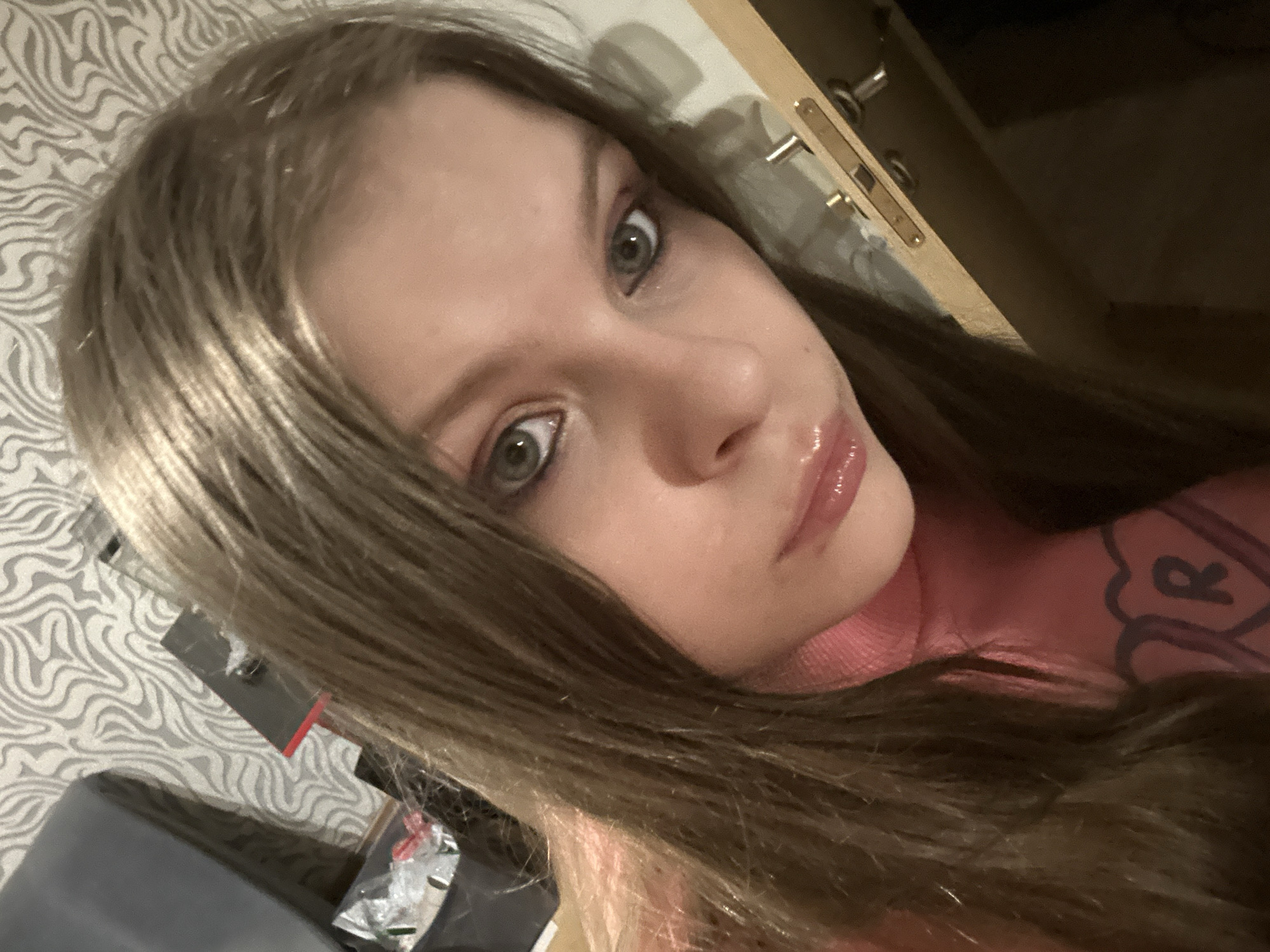 A fair-skinned person with long, light brown hair is posing for a close-up selfie. The individual has light-colored eyes and slightly glossy lips. They are wearing a pink top with a design on the upper right side. The background is complex, with a patterned wallpaper visible on the left side of the frame, while a wooden cabinet or door with metal hardware is seen at the top right. A dark object, possibly a chair, is situated in the lower left corner. There is nothing to indicate this photo was taken in Moscow, Russia.