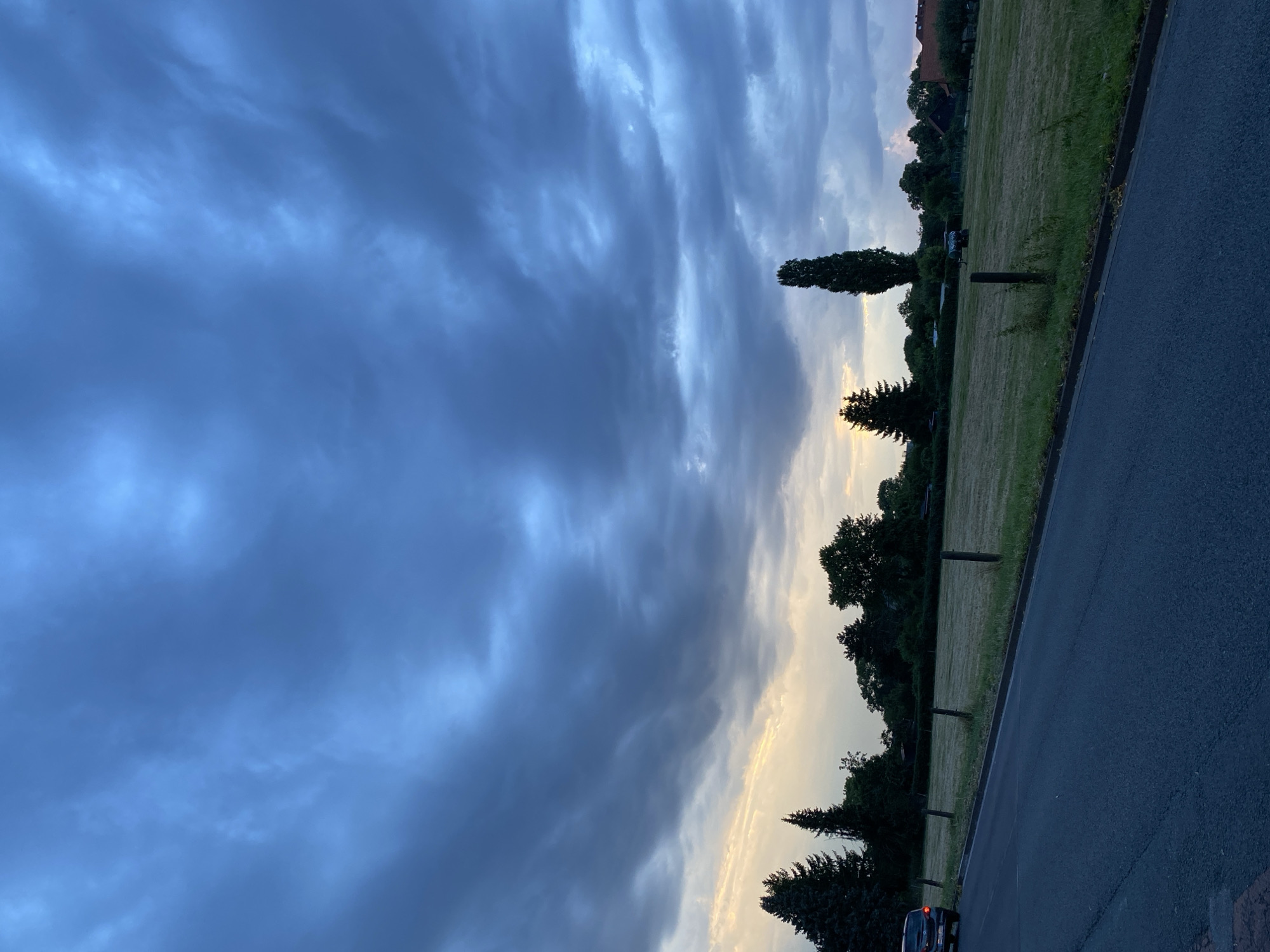 This image captures a dusk scene on a street in Rinteln, Germany, likely in a rural or suburban area. The sky is dominated by dramatic, dark blue and grey clouds, with streaks of pale yellow and white light breaking through near the horizon, suggesting either sunset or sunrise.
In the foreground, a dark asphalt road curves from the bottom left of the frame into the distance. A dark-colored car is visible on the left edge of the road. The road is bordered by a grassy verge and a low fence made of wooden posts.
Beyond the road is a large, open field of dry grass, possibly a meadow that has been recently mown. Several wooden fence posts are scattered across the field. In the middle ground, a variety of trees form a dense line across the landscape, including several tall evergreen trees on the left and a very tall, slender deciduous tree in the center. Further in the background, hints of residential buildings with red roofs can be seen peeking through the trees. The overall atmosphere is moody and atmospheric, with the gathering clouds indicating a potential change in weather.