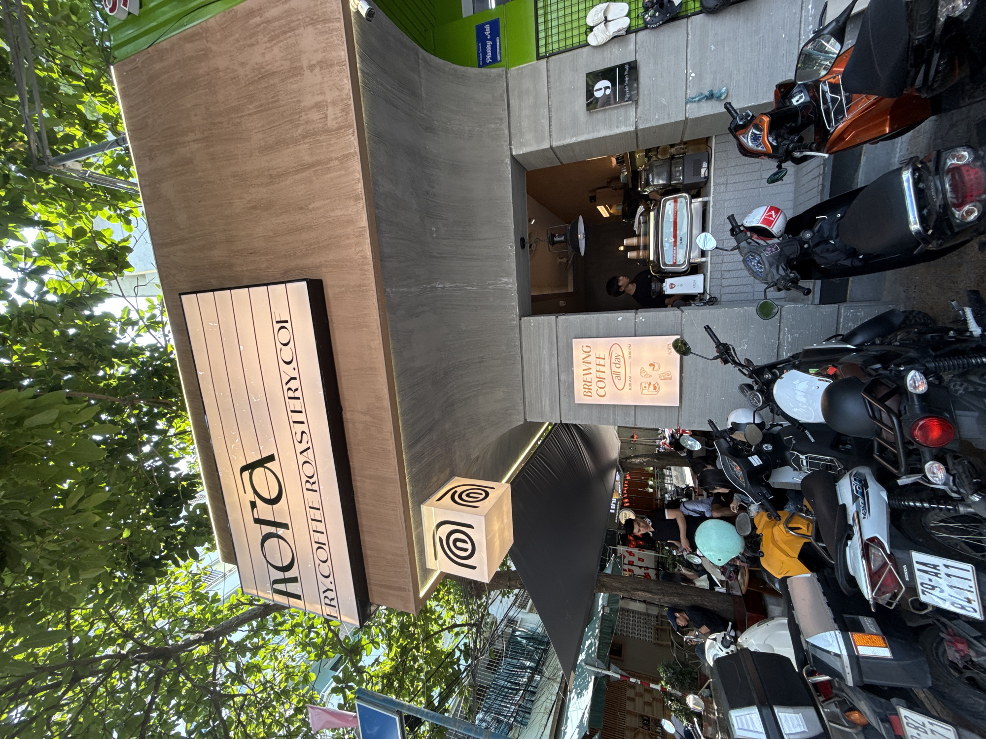 This eye-level, vertical shot depicts the vibrant street entrance of the "nora" coffee roastery in Nha Trang, Vietnam, on a bright, clear day. The coffee shop facade combines a warm, wood-textured panel on the left with a modern, concrete-like archway and vibrant green accents above. Lush green tree foliage shades the upper left portion of the image.
Prominently featured is a white, illuminated sign displaying "nora" in large black letters, with "ERY.COFFEE ROASTERY.COF" beneath it. A distinctive white cube-shaped light fixture with a spiral logo hangs near the entrance. Inside, a barista is visible behind a coffee machine, serving customers from a small, minimalist counter. Another illuminated sign details "BREWING COFFEE all day" from "6:30 AM - 10:00 PM."
The foreground and right side of the image are filled with numerous parked motorbikes and scooters, indicating a bustling urban environment. Several people are scattered around the vehicles, some wearing helmets, suggesting activity typical of a city street. Details like "HONDA" branding and a license plate reading "79-AA 841.11" are visible on the scooters. Additional signs with Vietnamese text, "Phương Anh" and "Nguyên Thiên Thuật," are also present above the entrance. The bright sunlight suggests it is daytime.