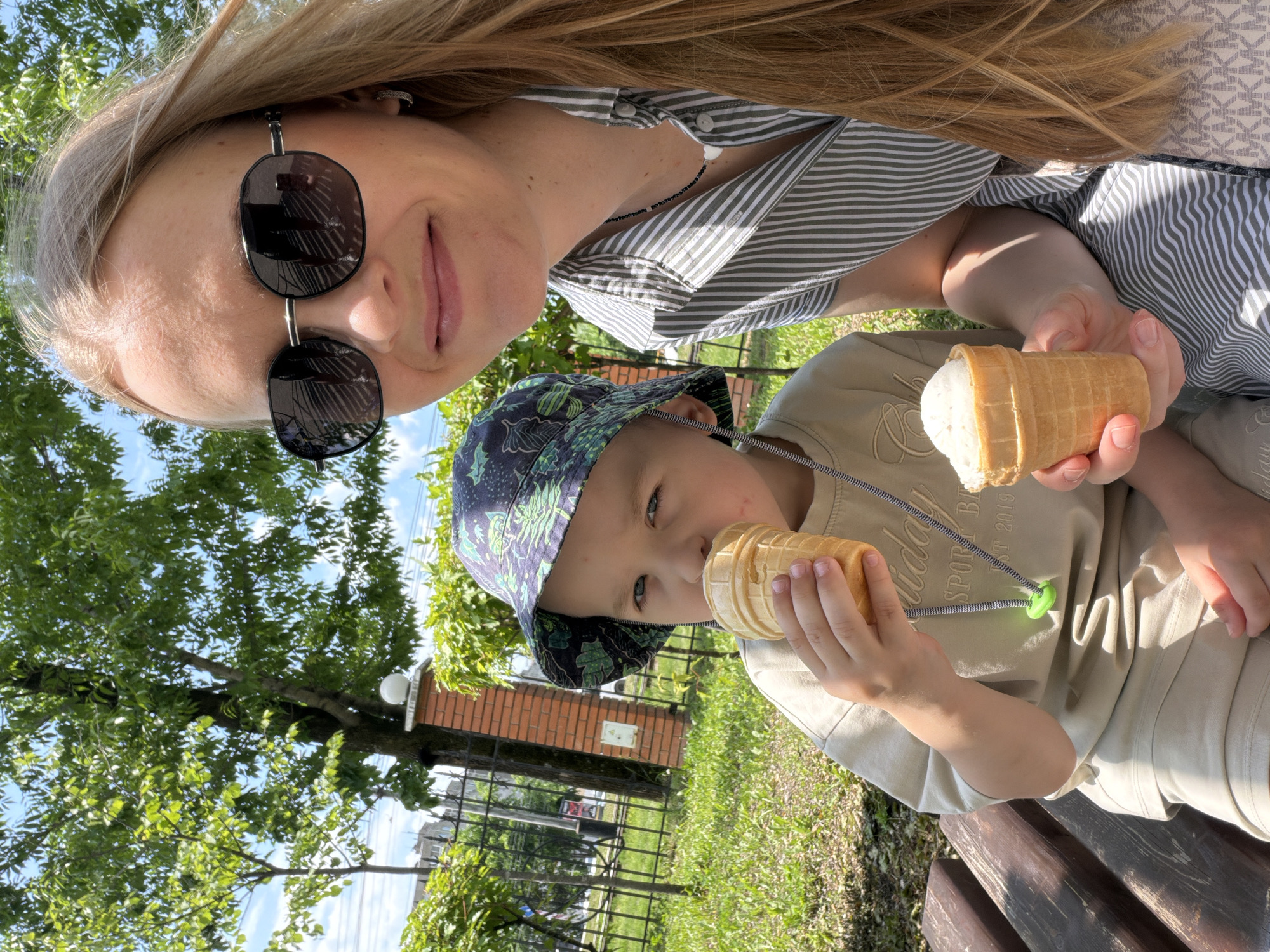 The image depicts a woman and a young boy sitting on a wooden bench outdoors, enjoying ice cream. The woman, with long blonde hair and wearing sunglasses, is on the right side of the frame, holding an ice cream cone. The boy, on the left, is wearing a patterned bucket hat and a light-colored t-shirt, and is also eating an ice cream cone. They are seated in what appears to be a park or public garden, with green trees and grass visible in the background. A brick pillar with a light fixture is present behind the boy. The overall setting suggests a warm, sunny day. There is no visible indication of the specific location within the city of Khabarovsk, Russia.