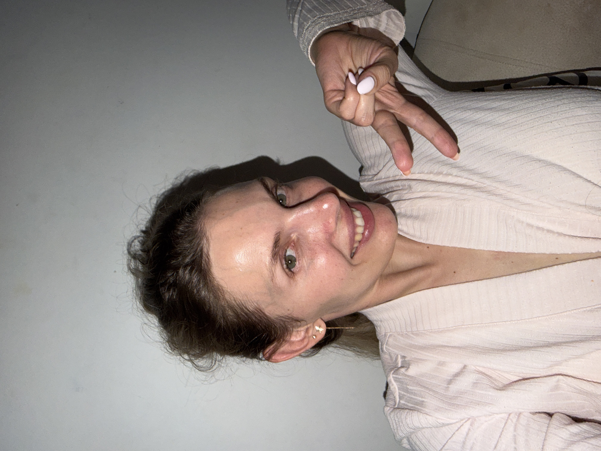 A woman is posing for a picture. She is wearing a light pink, ribbed top and smiling with her teeth showing. Her hair is brown and wavy, and she is wearing earrings. Her left hand is raised, making a peace sign. The background is a plain wall, and a chair is visible in the upper right corner. The location in Budva, Montenegro, is not evident from the image.