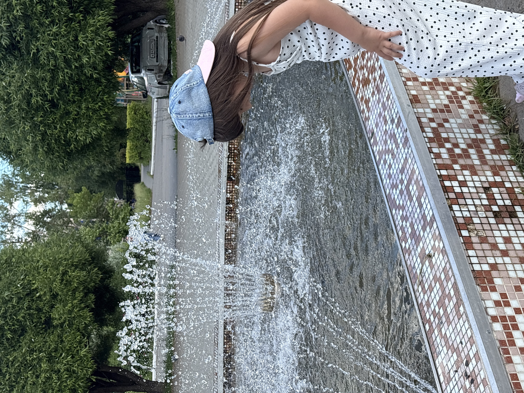 A young girl, wearing a denim baseball cap and a white dress with black polka dots, leans over the edge of a fountain, her face close to the water. Her hair, long and brown, cascades over her shoulders. The fountain is active, with streams of water spraying upwards and outwards, creating a misty spray. The fountain's edge is decorated with patterned tiles, a mix of brown and white squares arranged in a diamond pattern. To the left of the fountain, lush green trees and foliage are visible, suggesting an outdoor setting. In the background, beyond the trees, a grey car is parked. The overall scene appears to be a bright day, possibly in the summer, as indicated by the greenery and the activity of the fountain. The girl's posture suggests curiosity or fascination with the water.