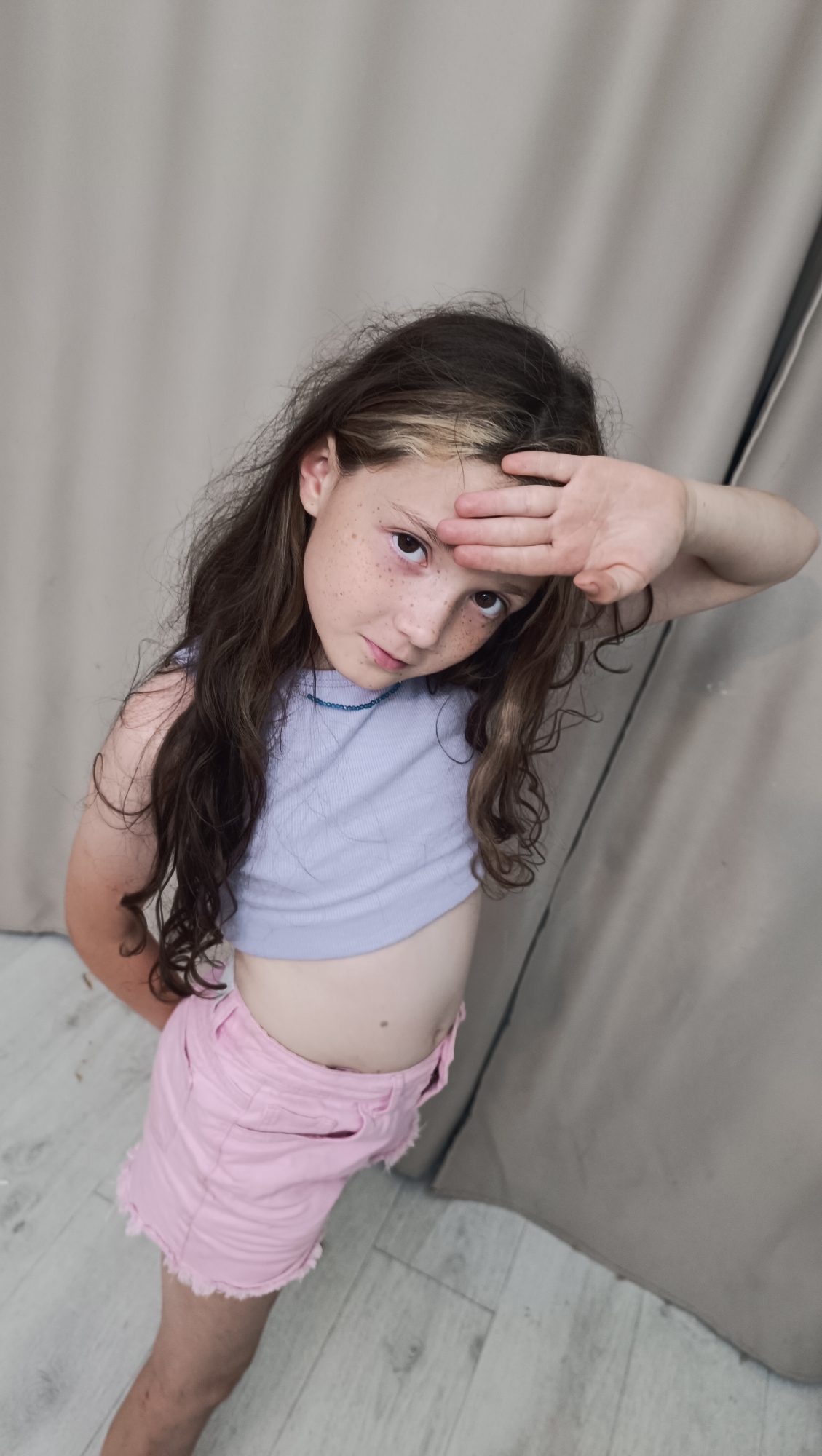 A young girl with long, wavy brown hair, some of which is dyed blonde, stands in front of a beige backdrop. She has light skin with freckles scattered across her nose and cheeks. The girl is wearing a light purple crop top and light pink denim shorts with frayed edges. She has a thin blue beaded necklace around her neck. Her right hand is raised to her forehead, shielding her eyes as she looks towards the camera. Her expression appears serious or perhaps thoughtful. The background consists of a light-colored, possibly wooden floor and a textured, neutral-colored curtain or wall. The lighting suggests it is daytime, and there are soft shadows on the backdrop. The girl is the sole subject in the image.
