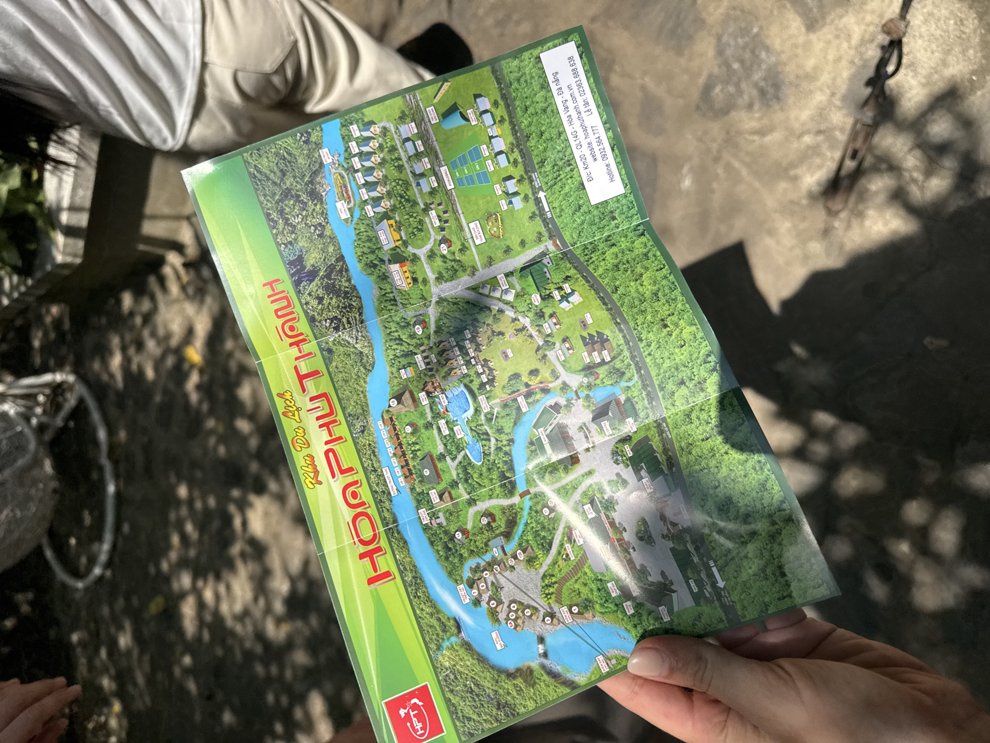 A person holds a folded map of Hoa Phu Thanh tourist resort, located in Hòa Vang district of Đà Nẵng city, Vietnam. The map details the layout of the resort with various attractions. A portion of a bicycle is visible in the background, along with shadows. The photo is taken outdoors.