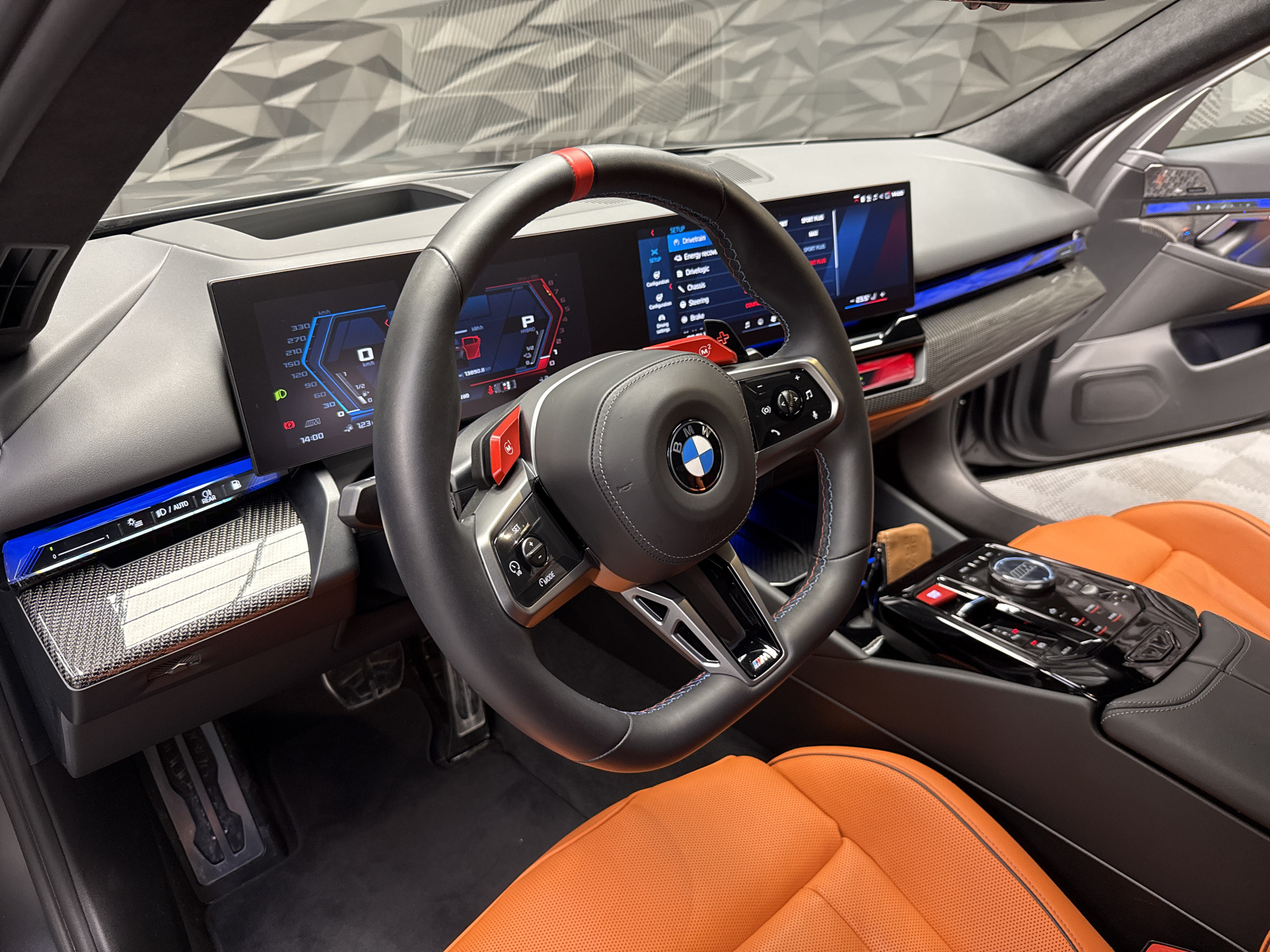This image shows the interior of a BMW car, focusing on the driver's cockpit. The steering wheel, prominently displayed, has a red stripe at the top and red M buttons. The dashboard features two digital screens, one behind the steering wheel displaying speed and other driving information, and another to the right displaying the car's settings menu, including options like "Drivetrain" and "Chassis." The car has orange leather seats and carbon fiber trim on the dashboard and door panels. The center console includes various buttons and controls. The lighting in the interior appears to be a mix of natural light and blue ambient lighting. There is no information provided in the image to indicate this scene is located in the city of Plovdiv, Bulgaria.