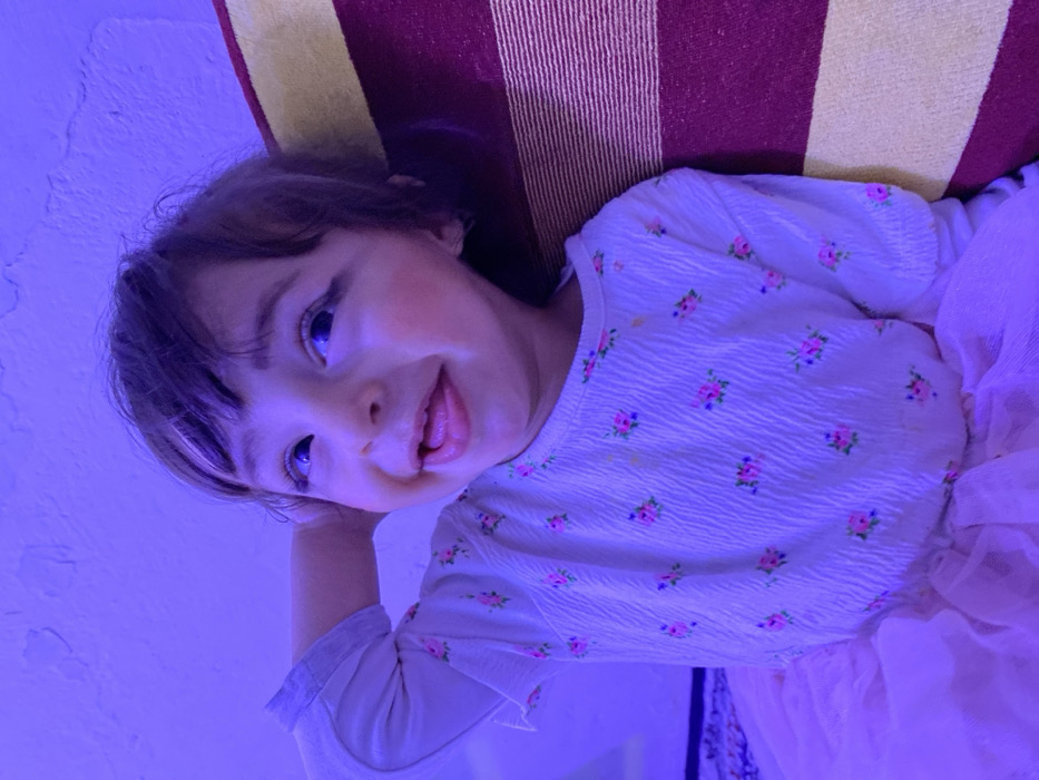 This image, taken indoors in the city of Kandahar, Afghanistan, features a young child under prominent blue-purple illumination. The child, with dark hair and bangs, is positioned centrally, looking upwards and slightly to the right, displaying a broad smile with the tongue partially extended. Black winged eyeliner is applied to the upper eyelids, and pink blush is visible on the cheeks. The child wears a long-sleeved white textured shirt adorned with small pink and green floral patterns, paired with a light pink tulle skirt. The child's left arm is bent, with the hand positioned behind the head. To the right, a cushion with alternating horizontal stripes of maroon and yellow is present. The background consists of a textured white wall, and a patterned rug is partially visible in the lower-left foreground.