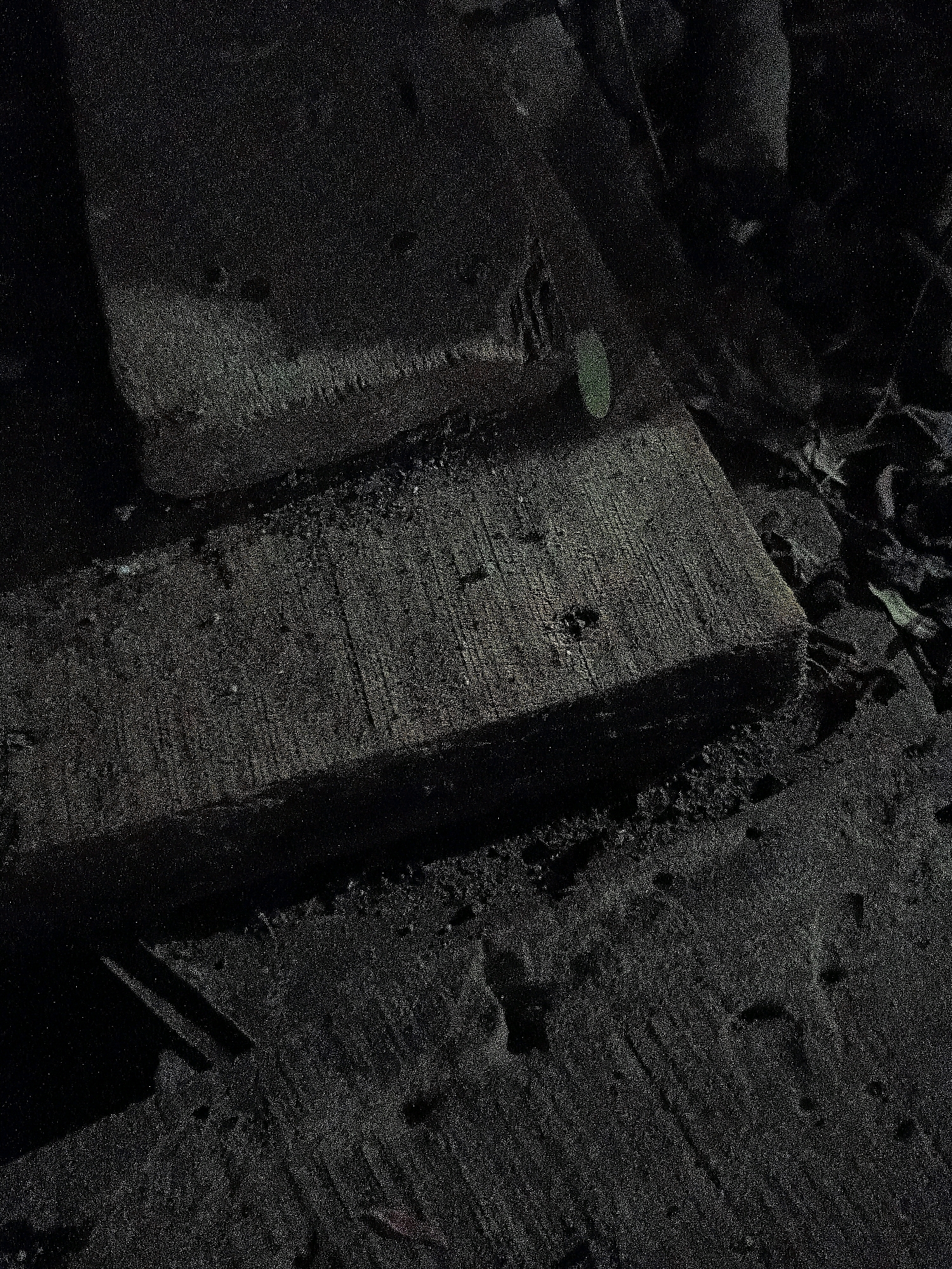 The image depicts a close-up, low-light view of rough, dark, textured surfaces that appear to be concrete or asphalt. There are two distinct horizontal planks or sections of material, one above the other, with a gap between them. The upper plank shows some texture and what could be dirt or debris. A small, round, greenish object, possibly a leaf or a piece of debris, is visible on the surface of the upper plank. The lower plank is wider and also exhibits a rough texture with visible grain lines. Small indentations or holes are scattered across its surface. The surrounding area is dark and appears to be an outdoor environment with some loose debris and foliage visible in the background. The lighting is dim, creating strong shadows and a grainy texture throughout the image. Without further context or identifying features, it is impossible to determine the specific location within the city of Cikal, Indonesia, or to identify any people, actions, or specific events.