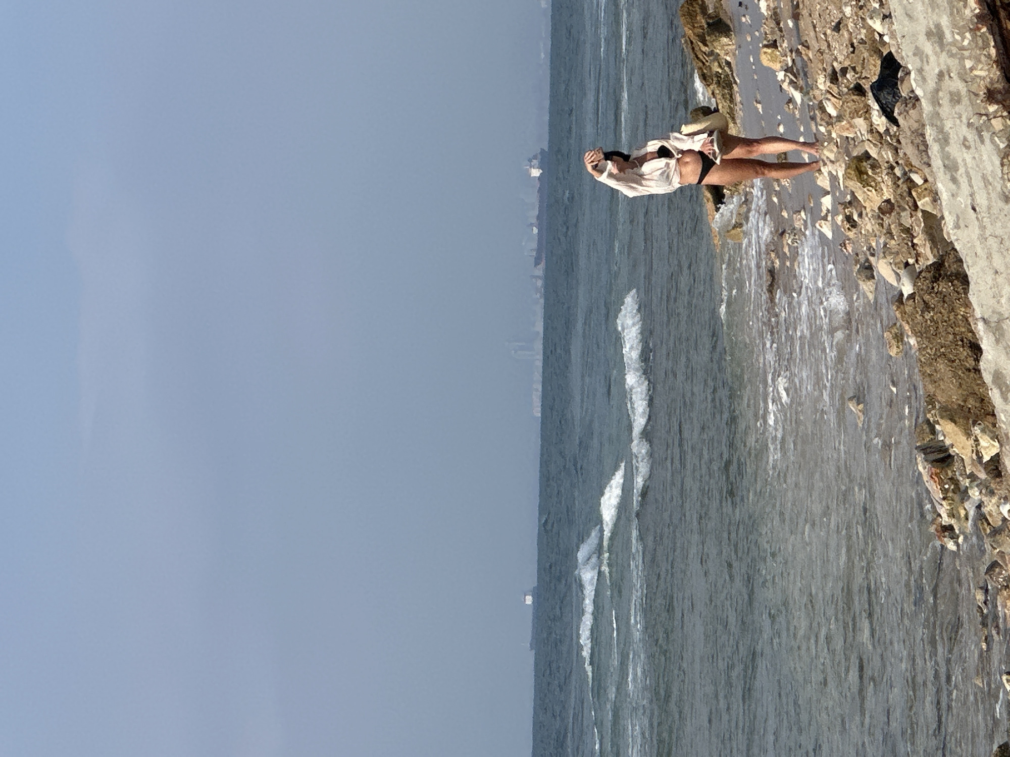 A woman stands on a rocky beach in Haifa, Israel, holding her phone up as if taking a selfie. She is wearing a white open shirt over a black bikini. The ocean is to her left, with gentle waves lapping at the shore. In the distance, several large cargo ships are visible on the horizon, and the hazy skyline of Haifa can be discerned. The sky is a pale blue, with some wispy clouds. The beach is covered in various sizes of rocks and pebbles. A bag is visible near the woman's feet.