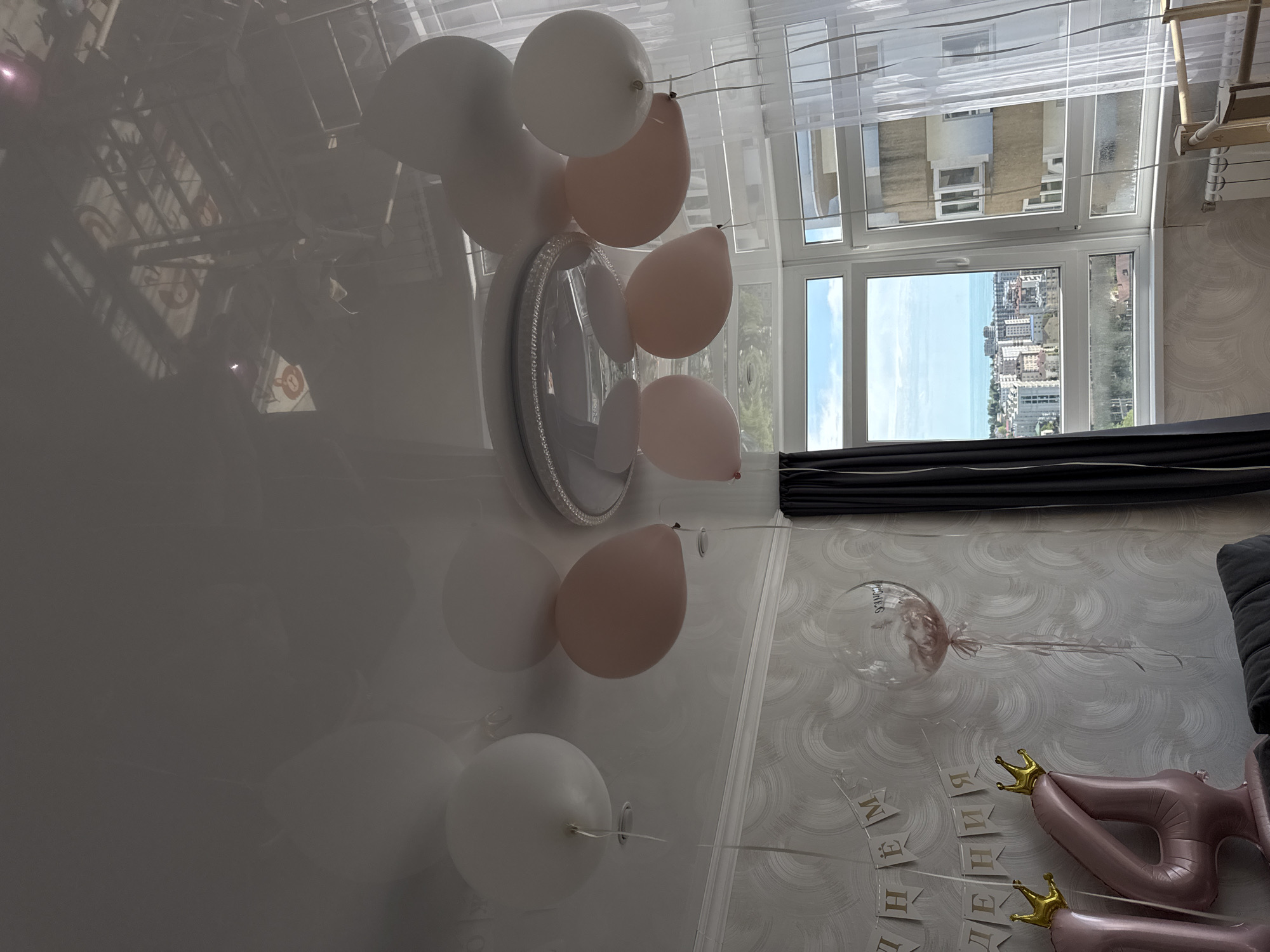 A room is decorated with balloons, some white and some pinkish, attached to the ceiling and wall. A decorative mirror is also mounted on the wall. A window provides a view of the city of Sochi, Russia, and the Black Sea. Part of a sofa is visible in the bottom right of the image. Additional decorations and balloons can be seen in the lower right area of the room.