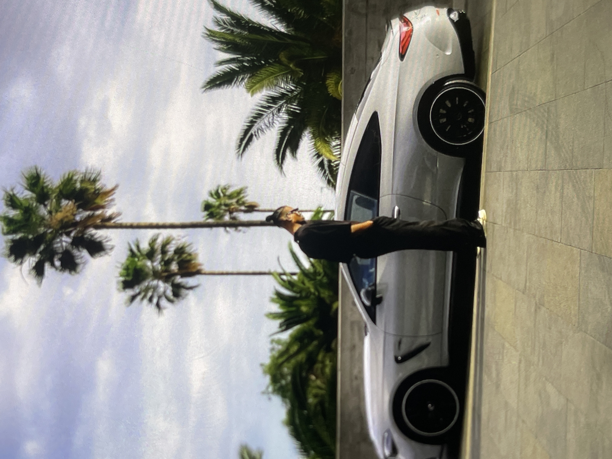 A person dressed in black stands beside a silver coupe parked on a tiled surface. The person has their back to the camera, with their hands in their pockets, and is looking down towards the car. The car is a two-door coupe with black rims and a sleek design. Behind the car and person, palm trees and lush green foliage are visible against a cloudy sky. The setting appears to be outdoors, possibly in a warm climate, with a sense of luxury and leisure.