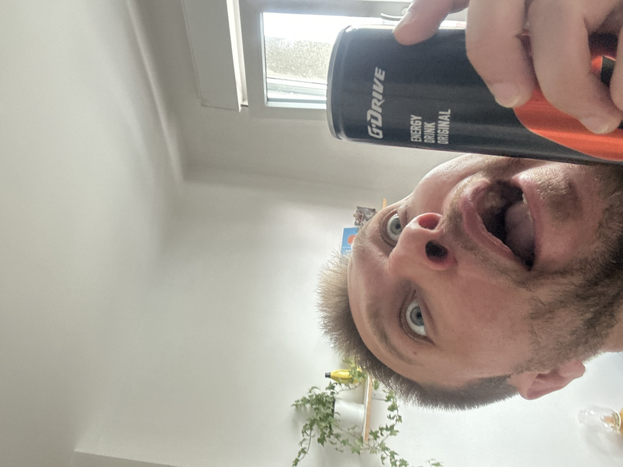 The media file shows a close-up, upside-down shot of a person's face as they hold a can of G'DRIVE energy drink. The person, who appears to be male with light skin and blue eyes, has an open mouth and wide eyes, suggesting surprise or an attempt to drink from the can. He has short, light brown hair and some stubble on his chin. The can is black with white text that reads "G'DRIVE ENERGY DRINK ORIGINAL". Part of an orange can or label is visible on the right side, behind the black can.
The background consists of a white ceiling and a window with what appears to be a textured or frosted glass, suggesting daylight. The overall setting seems to be indoors, possibly a home or office, with a glimpse of a white wall and some greenery from a plant in the lower left corner. There is also a hint of other objects on a shelf in the background, including what looks like a banana. The lighting is bright, indicating it's daytime. The orientation of the image is unusual, with the person's head at the bottom and the can at the top, as if the photo was taken from a low angle looking upwards.