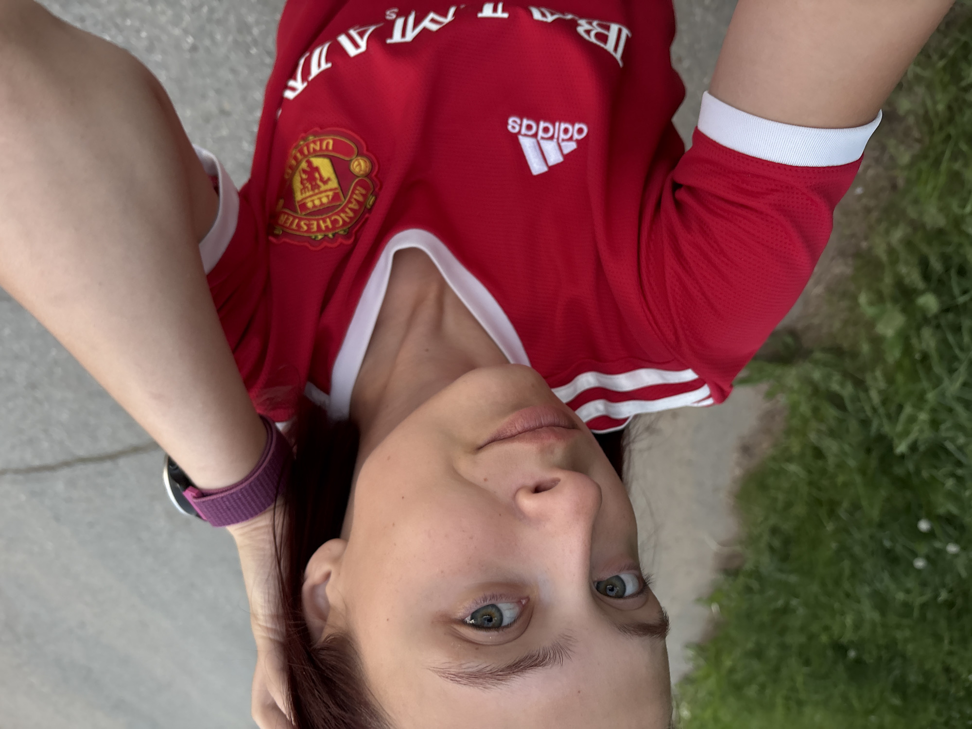 A fair-skinned woman is wearing a red Manchester United Adidas jersey. She has a fitness tracker on her left wrist. The surrounding environment appears to be an outdoor area with a concrete ground and some greenery. The image's location details (Прикамский, Russia) are not visually discernible from the photo.