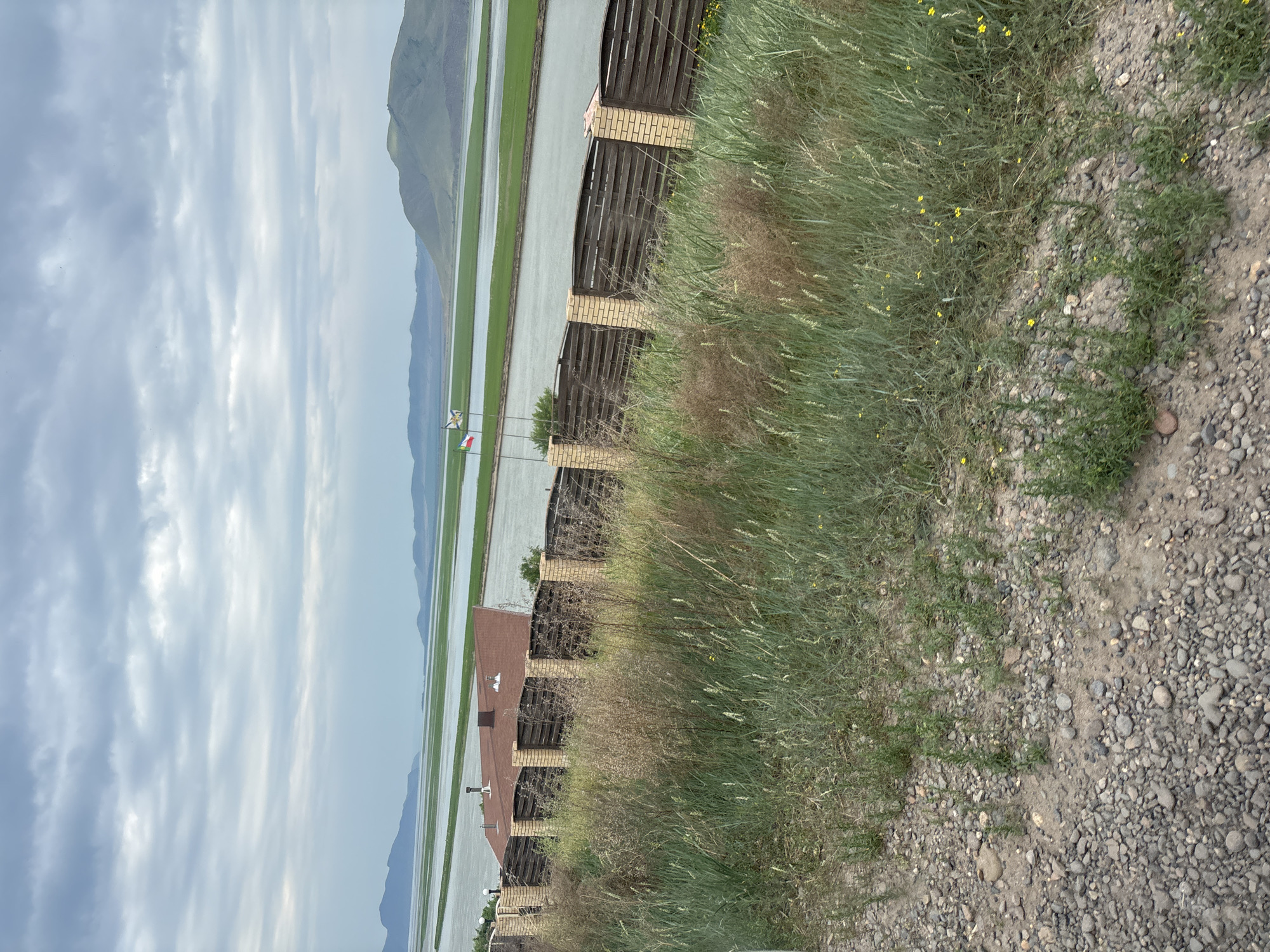 The image depicts a scenic view of a body of water, likely a river or lake, with verdant green fields stretching out in the distance. Rolling hills and mountains form the backdrop under a cloudy sky. In the foreground, a low-rise building with a brown roof is visible, bordered by a fence constructed with brick pillars and dark wooden slats. Tall, dry grasses and some yellow wildflowers grow in front of the fence, with a gravelly path in the immediate foreground. Two flags are planted near the fence, one with a blue and white stripe pattern and another with red, white, and blue stripes. The scene appears to be in a rural or semi-rural setting within the city of Ust-Abakan, Russia. There are no people visible in the image.