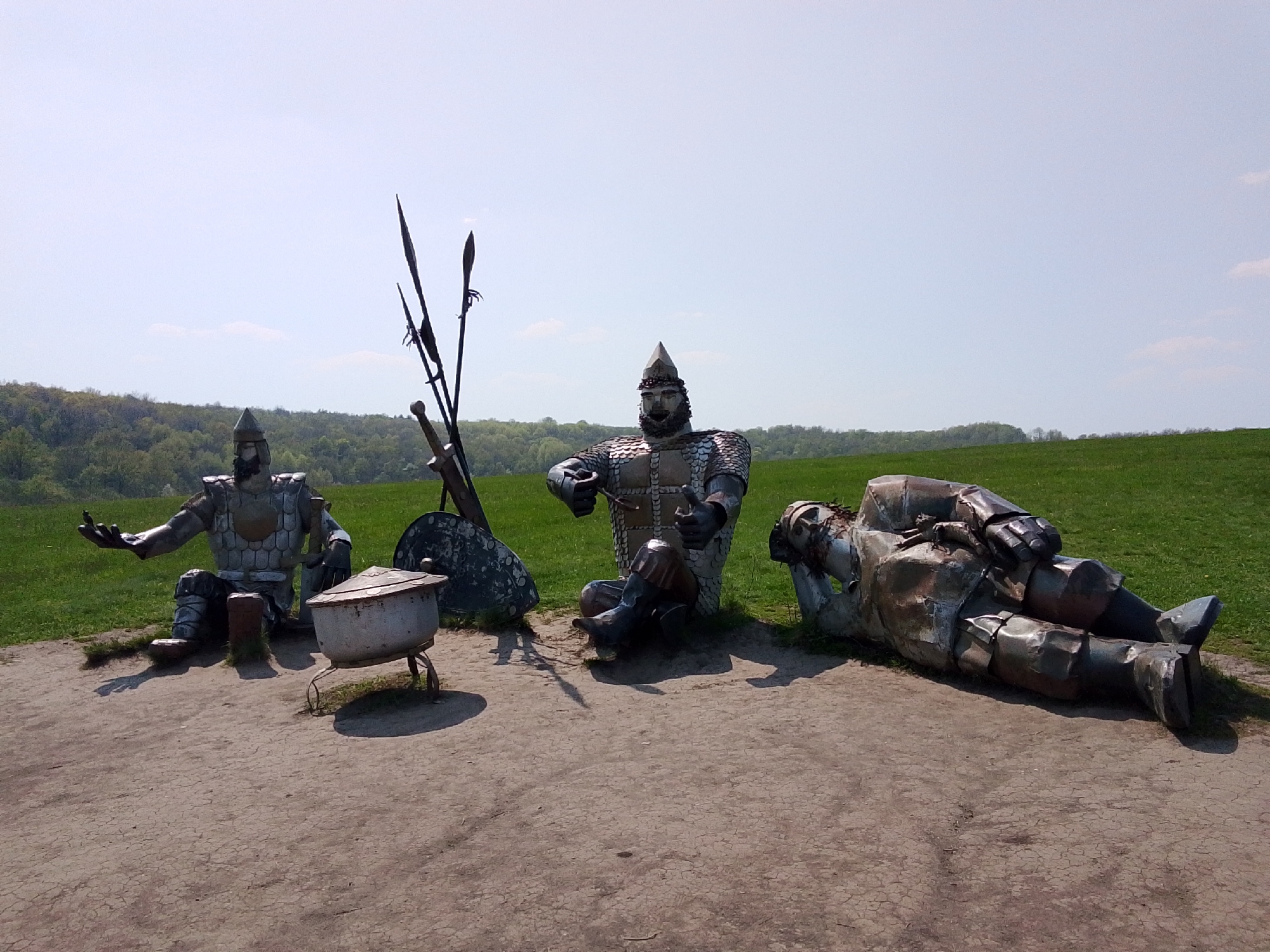 The image shows a landscape with three metal sculptures of medieval warriors on a patch of bare ground, possibly dirt or sand. Behind them is a grassy hill and a line of trees in the distance, under a clear, light blue sky.
The leftmost warrior sits with his legs crossed, one arm outstretched with an open palm. Next to him is a small, metal pot on a three-legged stand. A shield, spears, and a sword lean against the pot.
The central warrior is seated with knees bent, holding a small object in his hands.
The rightmost warrior lies on his back, seemingly resting or deceased.
The scene is located in Каменка, Russia.