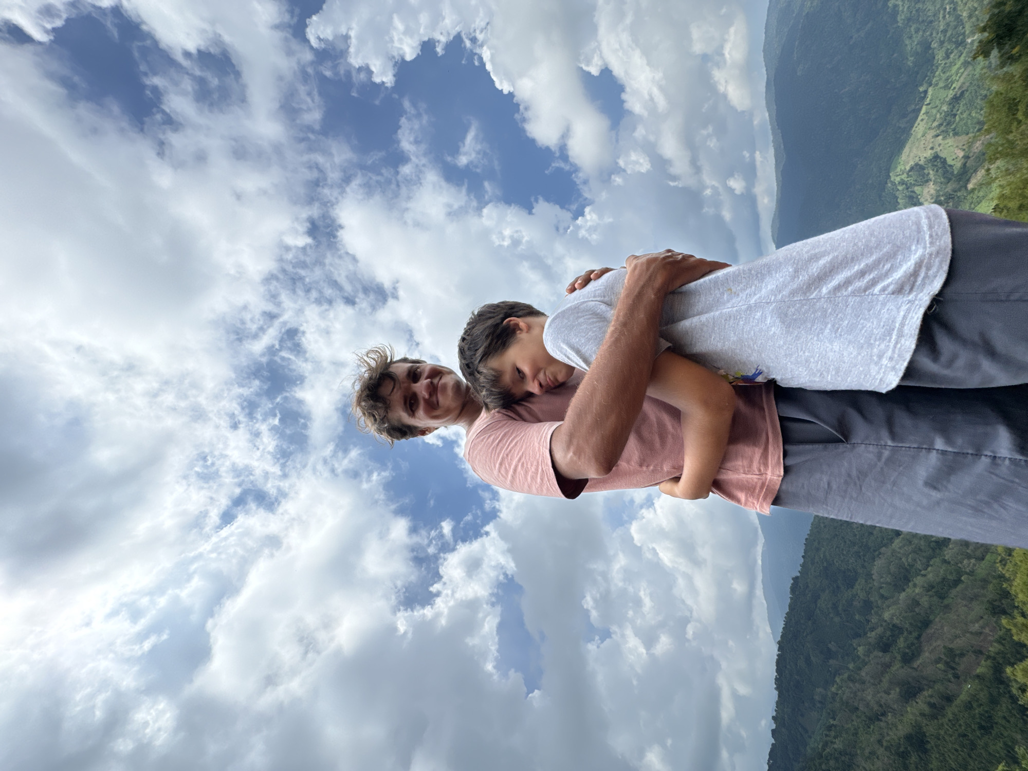 Two males, one adult and one child, are embracing each other outdoors. The adult, on the left, is wearing a pink t-shirt and grey pants. The child, on the right, is wearing a grey t-shirt. They are standing on a hill with a background of lush green mountains and a cloudy sky. The image is taken from a low-angle perspective. The location appears to be Nao Sẻ, Vietnam, a mountainous region known for its natural beauty.