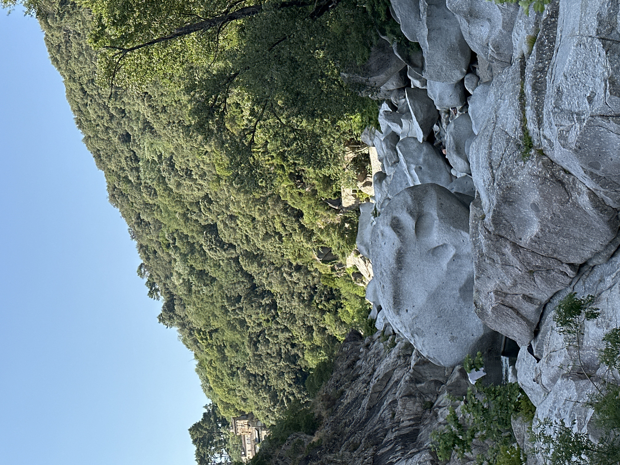 The image displays a steep, densely forested mountainside adjacent to a rocky, uneven terrain with large boulders. A clear blue sky dominates the upper left portion of the frame. The foliage on the mountain appears lush and green, suggesting a temperate climate and likely daytime. Sunlight casts shadows on the rocks, highlighting their varied textures and shapes. One large, smoothed boulder in the foreground has several circular indentations, resembling a face. A small structure with a pale facade and multiple windows is visible at the base of the mountain on the left side, partially obscured by trees. The overall scene evokes a sense of natural beauty and rugged landscape. The orientation of the image is such that the sky is on the left and the terrain is on the right, suggesting the photograph was taken looking upwards or at an angle from a lower vantage point.