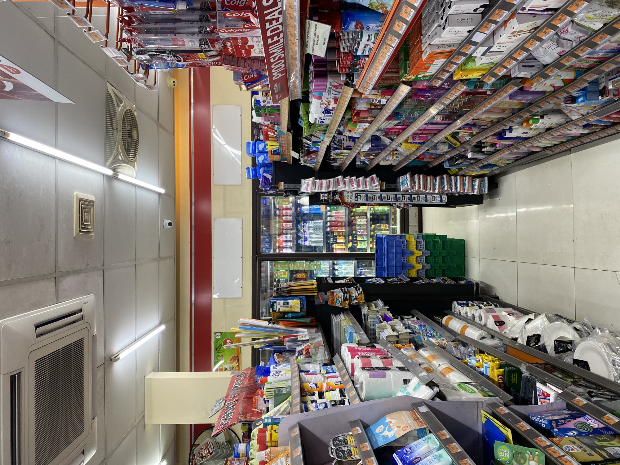 The image depicts the interior of a convenience store, likely a 7-Eleven, in Davao City, Philippines. The aisle features shelves stocked with various consumer goods, including toiletries, cleaning supplies, food items, and beverages. In the foreground, shelves on the left display items like facial tissue, paper towels, bottled water, and packaged snacks. To the right, shelves are filled with an assortment of personal care products, such as shampoos, conditioners, lotions, soaps, and toothpaste. Behind these shelves, refrigerated display cases contain drinks. The store is brightly lit by fluorescent ceiling lights and illuminated by advertising displays. Blue and yellow plastic crates are stacked near the end of an aisle. The overall impression is that of a well-organized retail environment, typical of a convenience store chain.