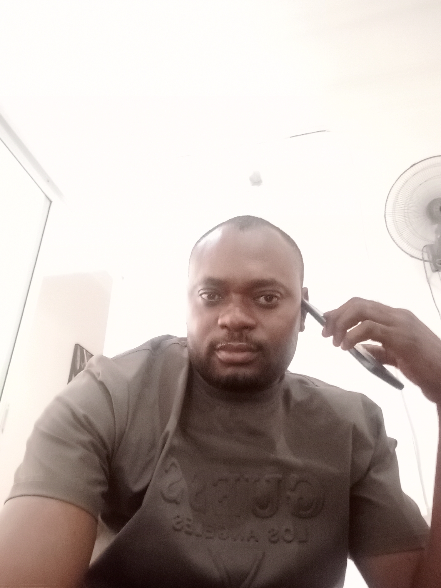 A medium-close-up shot shows a dark-skinned man with a shaved head holding a black smartphone to his left ear. He is wearing a grey t-shirt with raised lettering on the front. The background is a bright, white indoor space. A window and a white wall can be seen in the upper left corner. A white electric fan is mounted on the wall on the upper right side of the frame. Location details are unavailable from the image.