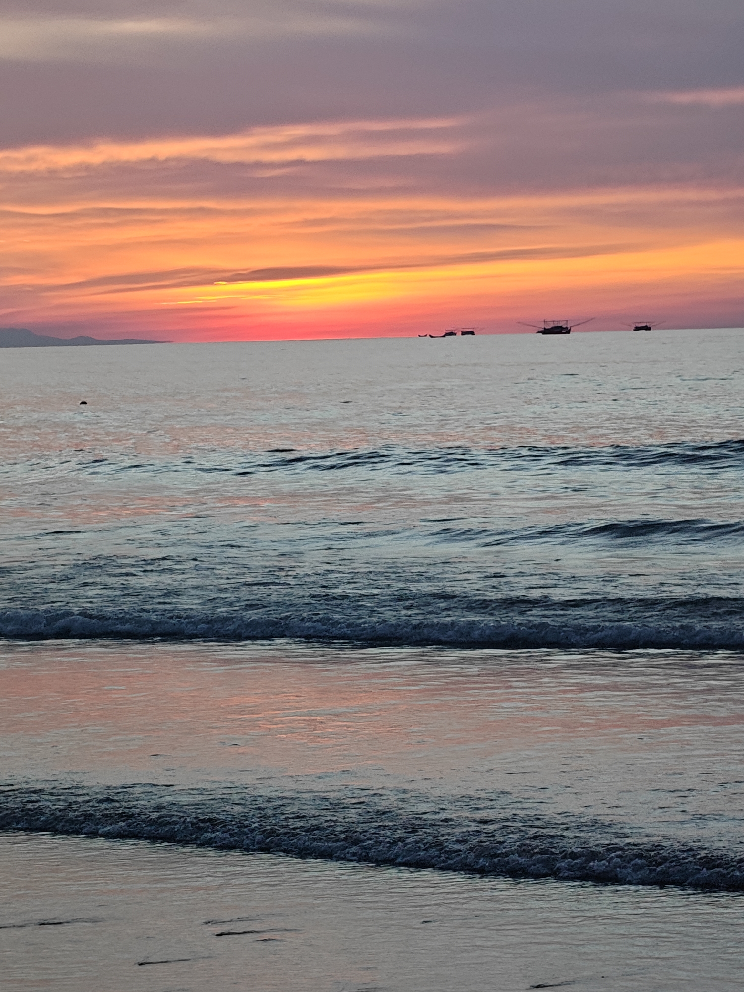 The image depicts a tranquil seascape at sunset, likely in Curehtunong, Indonesia. The sky is painted with vibrant hues of orange, yellow, and pink, transitioning to a soft purple at the top, characteristic of a tropical sunset. Several fishing boats are silhouetted against the horizon, suggesting their return to shore after a day's work. The sea is relatively calm, with gentle waves lapping at the shore. The water reflects the colors of the sunset, creating a shimmering effect. The foreground shows the wet sand of the beach, with the edge of a wave breaking. The overall atmosphere is peaceful and serene.