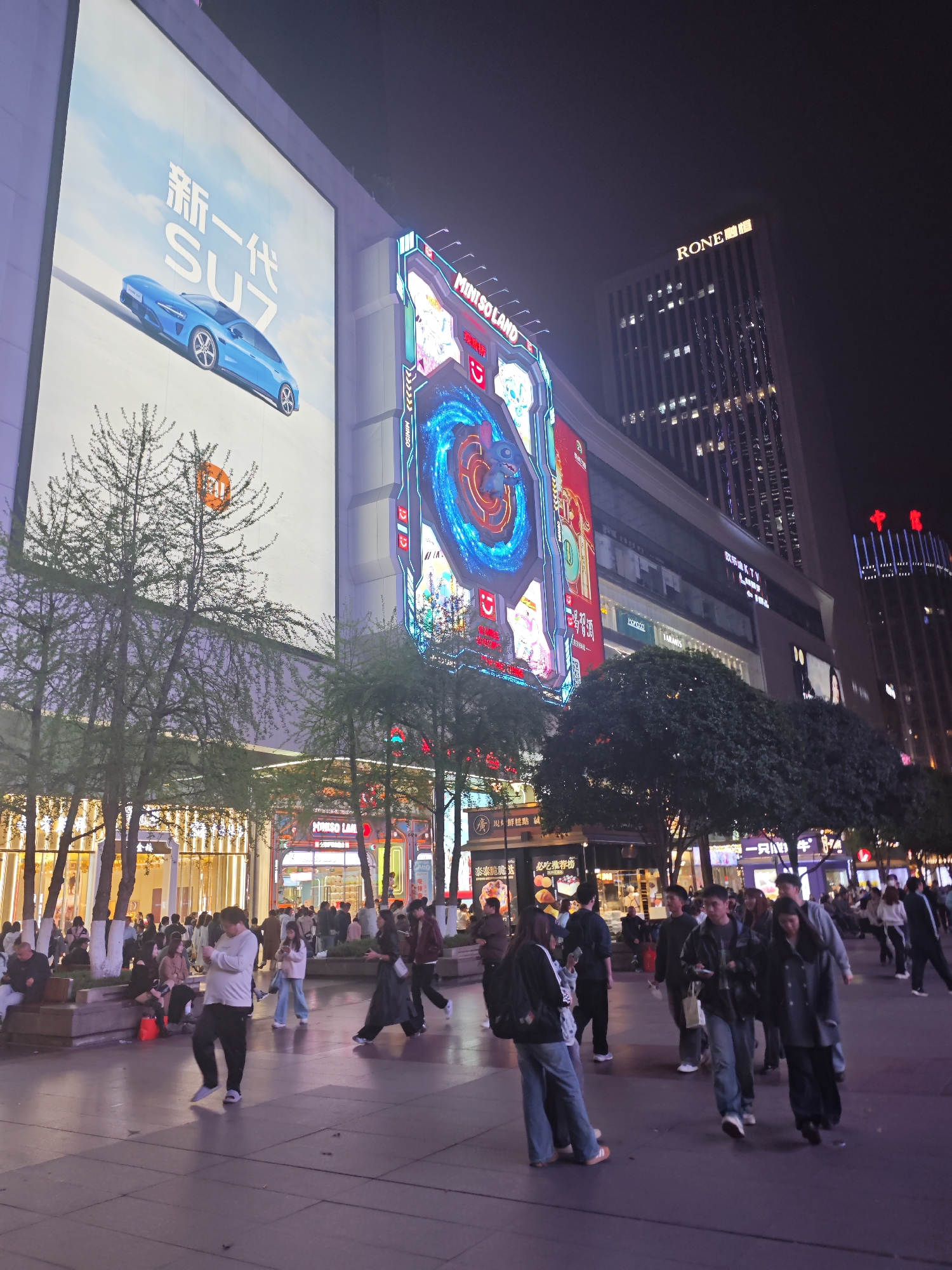 The photograph captures a bustling commercial district in Liangjiang, China, at night. Pedestrians populate a wide, paved plaza, some strolling, while others are seated on low benches. Illuminated buildings line the perimeter, featuring bright LED billboards and advertisements. A large billboard showcases a blue car and Chinese text, while another displays a colorful, animated MINISO advertisement. A tall building with the name "RONE" on top looms in the background, next to another tall building featuring a red emblem at the top. Trees are dispersed throughout the area, adding a natural element to the urban setting. The overall lighting is bright and colorful, reflecting off the pavement and creating a vibrant atmosphere.