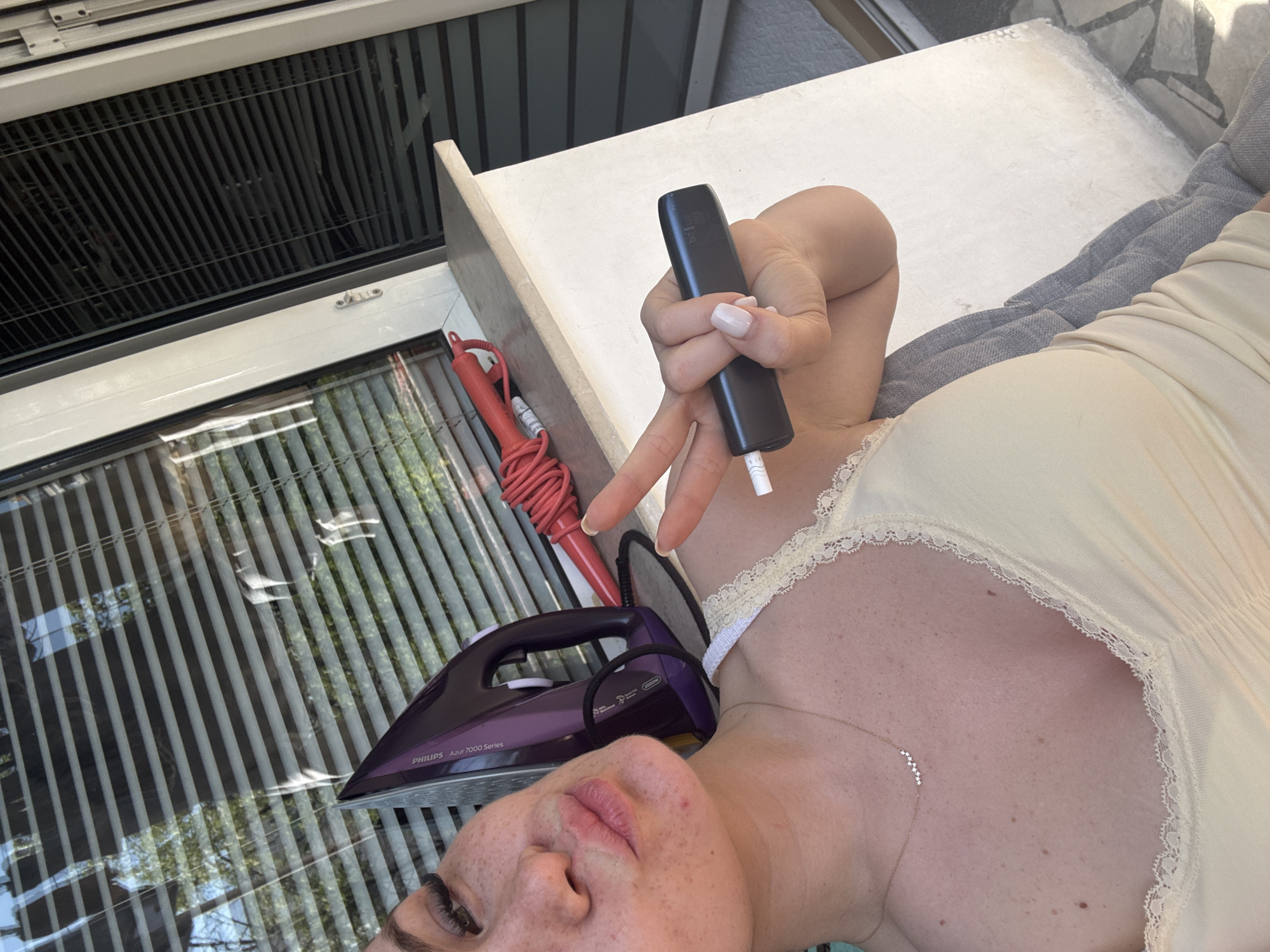 A selfie showing a woman with fair skin, freckles, and brown eyebrows, holding a dark gray electronic device between her fingers, next to her face and upper chest. The device is producing what appears to be a smoke-like vapor. She is wearing a light-colored, low-cut garment with a lace trim. Next to her is a purple Philips Azur 7000 series steam iron with a red cord. The background features a window with closed Venetian blinds, reflecting outdoor light. The location appears to be indoors, possibly in Plovdiv, Bulgaria, based on the European electrical outlet configuration of the iron cord. There is also a textured white surface nearby.