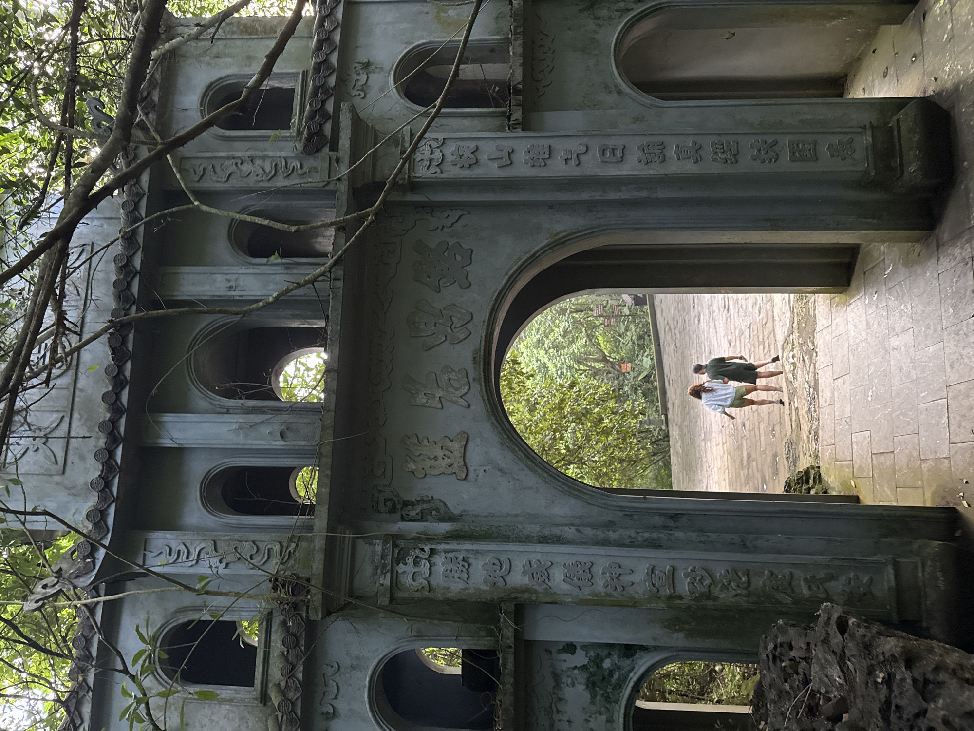 The image shows an ornate, weathered stone gate in Tây Hoa Lư Ward, Vietnam, partially obscured by dense foliage. The gate is a multi-tiered structure with arched openings and decorative carvings, including vertical inscriptions in Vietnamese. In the background, through the central archway, two individuals are walking away from the camera on a stone pathway. The environment appears to be a tropical or subtropical region, with lush greenery surrounding the stone structure. The stone itself shows signs of age and weathering, with moss and discoloration visible in places. The light suggests it is daytime, with sunlight filtering through the trees.
