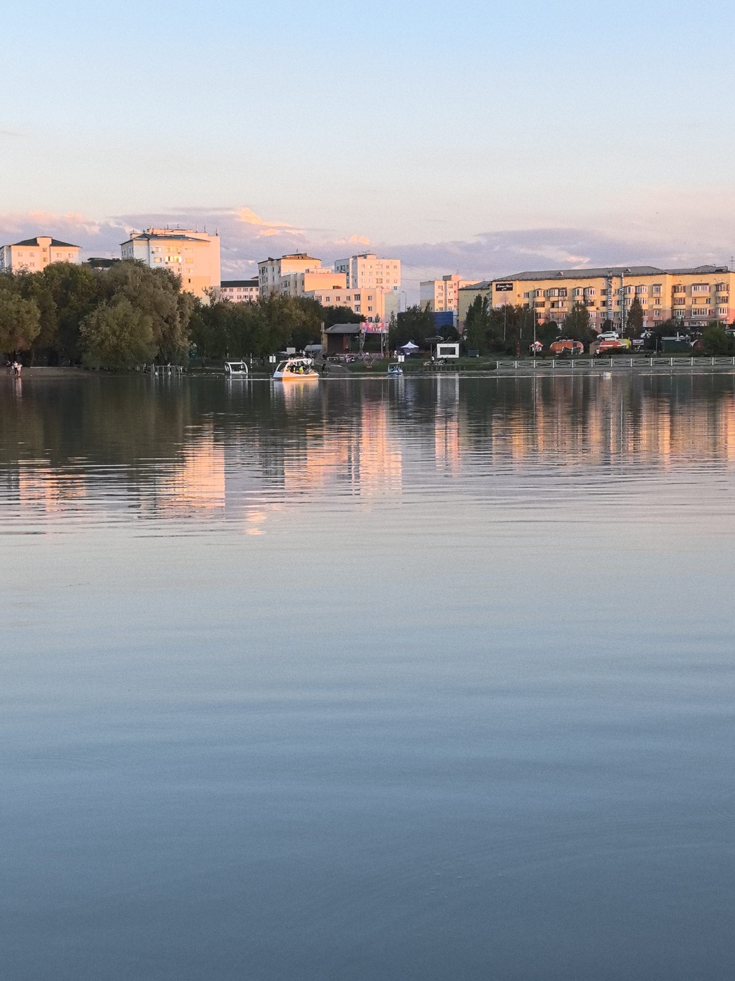 This is a photograph of the city of Taldykorgan, Kazakhstan, taken from across a body of water, likely a lake or a wide river. The image captures a serene late afternoon or early evening scene, with the setting sun casting a warm, soft glow on the buildings and clouds.
In the background, a row of apartment buildings lines the waterfront. These buildings vary in height and architectural style, with some appearing to be older Soviet-era structures and others more modern. They are painted in a range of pastel colors, including white, yellow, and pink, which are illuminated by the golden light. Trees and greenery are interspersed between the buildings and along the shoreline, creating a lush, park-like atmosphere.
On the water, several small boats are visible. One is a white, modern-looking vessel, and another is a yellow and white boat with people on board, suggesting it is a tour boat or a personal craft. The water itself is calm, with gentle ripples that reflect the colors of the sky and the buildings. The lower half of the image is dominated by the placid water, with soft blue and grey tones, indicating a clear and peaceful environment.
The sky above is a pale blue, with streaks of pink and orange clouds, further emphasizing the time of day. The overall impression is one of tranquility and urban beauty integrated with natural elements.