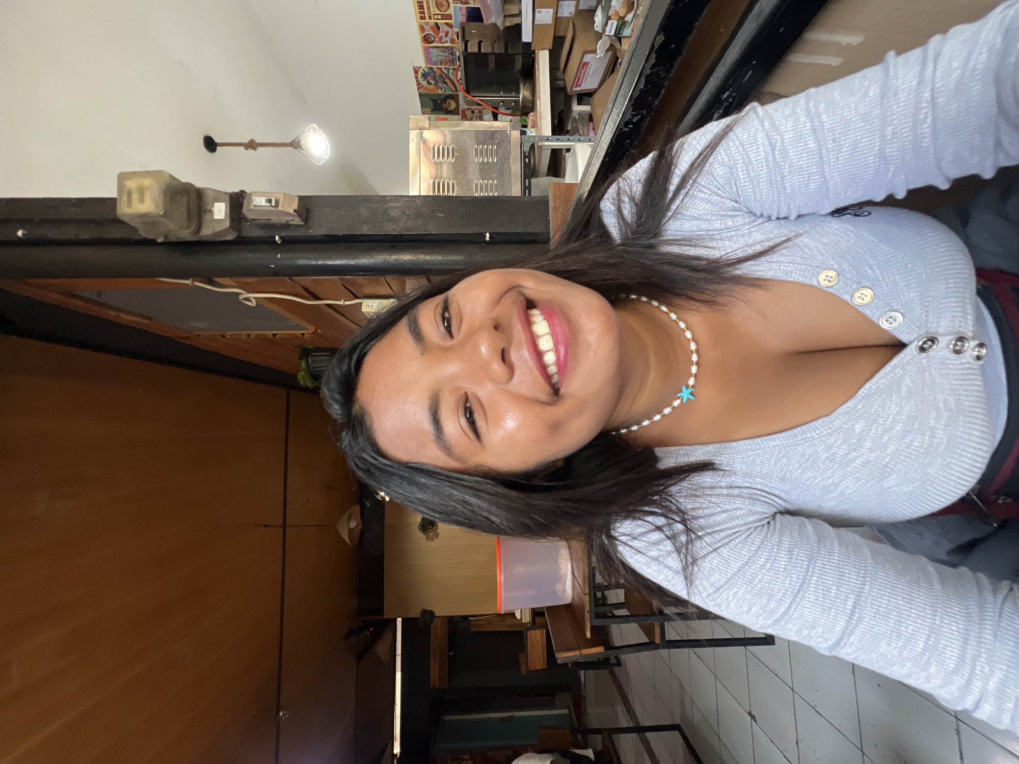 In a casual indoor setting, likely a cafe or restaurant in Kuta, Indonesia, a young woman with dark hair smiles at the camera. She is wearing a light gray, long-sleeved, V-neck top with buttons, and a pearl necklace with a turquoise star pendant. The background reveals wooden paneled walls, a metal pillar, and part of a kitchen area with appliances and shelving. Seating with wooden benches and tables are visible, along with a large translucent plastic container on a table. The lighting suggests it is daytime.