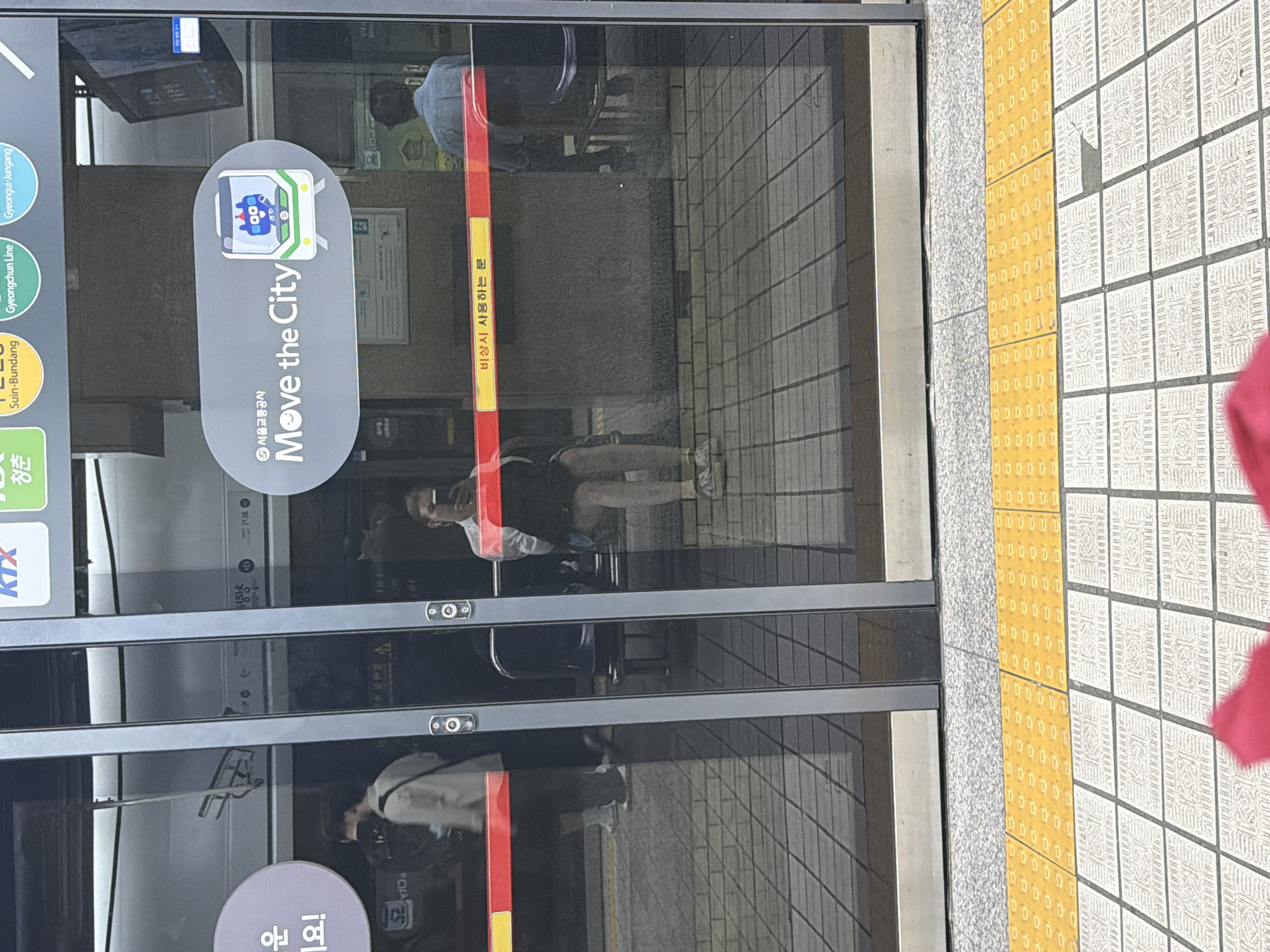 The image shows the interior of a subway train car in Seoul, South Korea, as seen through the glass doors. On the left, a person wearing a light-colored jacket and dark pants is walking away from the camera. In the center, a person with long hair is sitting on a bench, holding a smartphone, and appears to be taking a selfie. To the right, another person is seated on a bench. The train car has metallic doors with visible handles and buttons. A sticker on the door displays the Seoul Metro logo and the slogan "Move the City" with an illustration of a cartoon train. A yellow strip with red text that reads "비상시 사용하는 문" (Door for emergency use) is visible across the doors. The floor of the train car is tiled. Outside the train, a yellow tactile paving strip is visible on the platform. The overall environment suggests a typical subway station in Seoul.