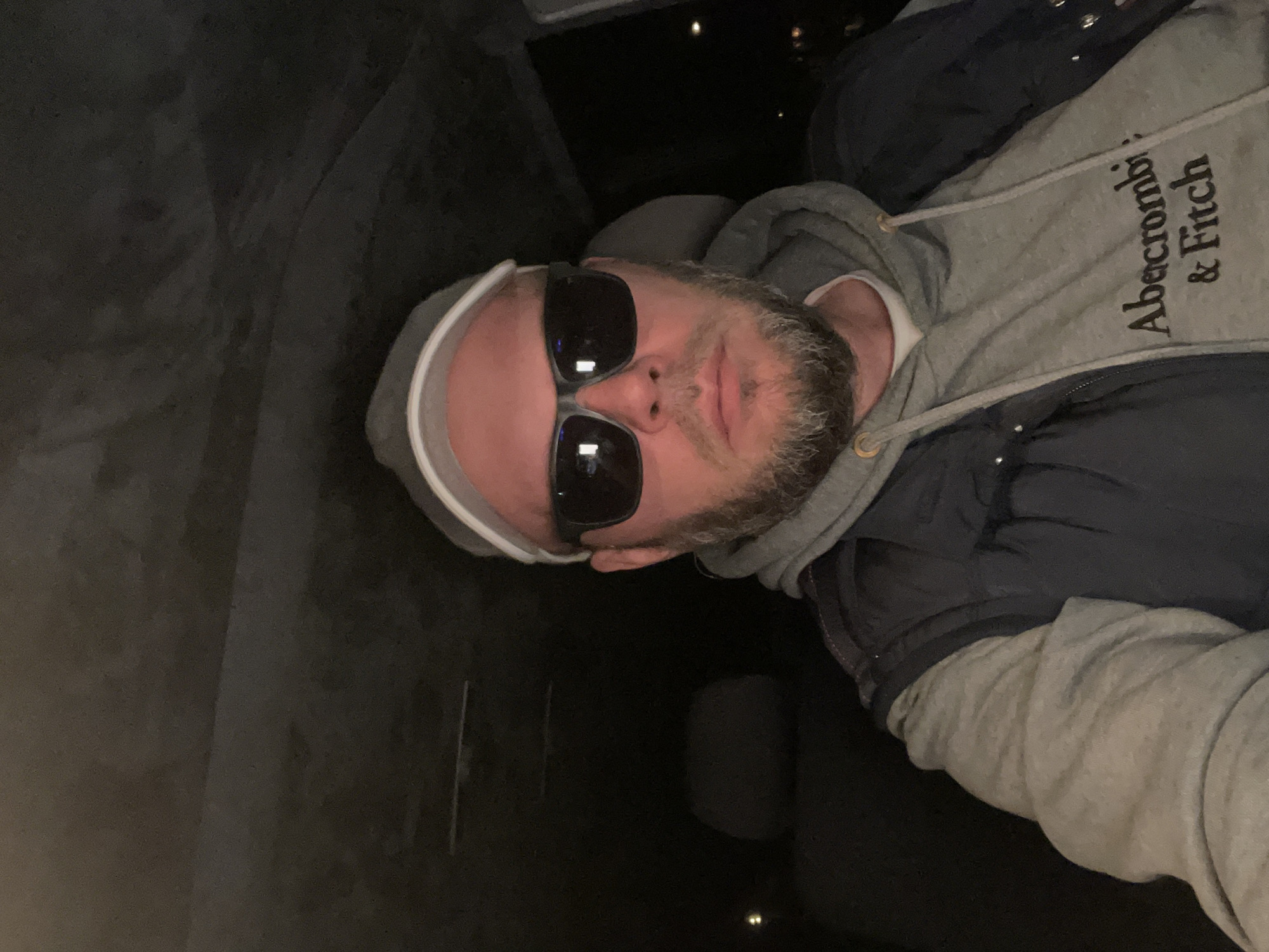 A man is depicted in a portrait-style photograph, taken from a slightly elevated angle. He appears to be reclining or lying down, looking directly towards the camera. The man has a beard and is wearing dark sunglasses, which obscure his eyes. He is dressed in a grey hooded sweatshirt (hoodie) with the hood pulled back, and a dark-colored jacket over it. A white band, possibly a headband or part of the hoodie's lining, is visible across his forehead.
The grey hoodie features the brand name "Abercrombi & Fitch" written vertically on the chest in dark lettering. The background of the image is largely dark and indistinct, suggesting an indoor environment or nighttime setting with low lighting. Reflections of what appear to be small light sources are visible in the lenses of his sunglasses. The overall mood is casual.
The scene takes place in Khimki, Russia.