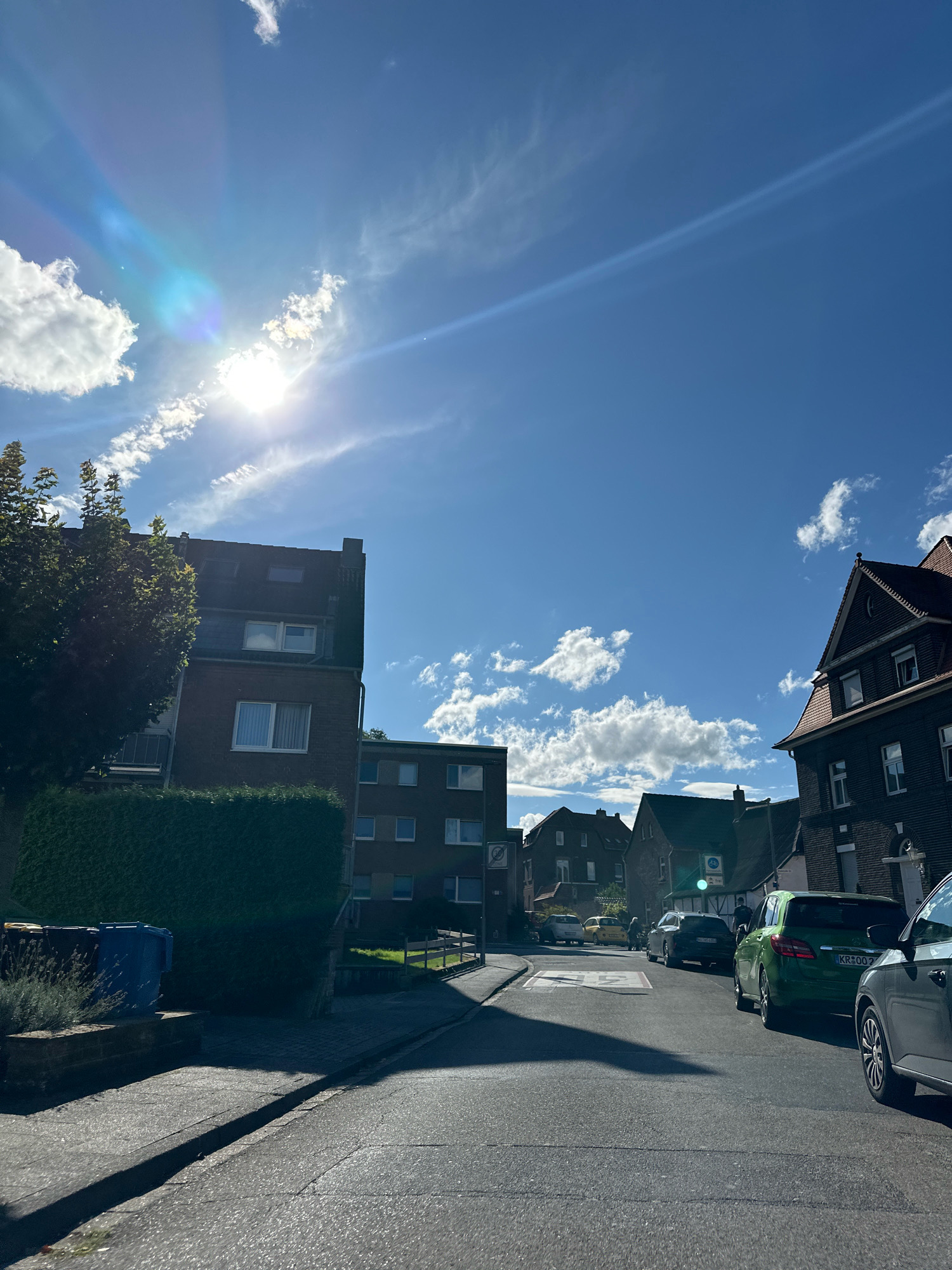 The image captures a sun-drenched street scene in Krefeld, Germany, likely on a residential street. The sky is a vibrant blue with scattered white clouds, and the sun is prominent, casting a lens flare with rainbow hues. The street is paved and appears to be a normal asphalt road.
On the left side of the street, a tall, multi-story brick building with multiple windows and a dark roof is partially visible, flanked by a neatly trimmed green hedge. Adjacent to the hedge are several large trash bins, including a blue one. Further down the street, other residential buildings can be seen.
The right side of the street features a dark-brick building with a red tiled roof, characterized by its distinctive gabled architecture and numerous windows. Several cars are parked along the street. A bright green hatchback is prominently featured in the mid-ground, and a dark-colored sedan is parked behind it. Another car, a grey SUV, is partially visible in the foreground on the right. In the distance, other vehicles and a few people are present.
The lighting suggests it is daytime, likely late afternoon or early evening, given the long shadows cast across the road. The overall impression is a quiet, ordinary day in a German city.