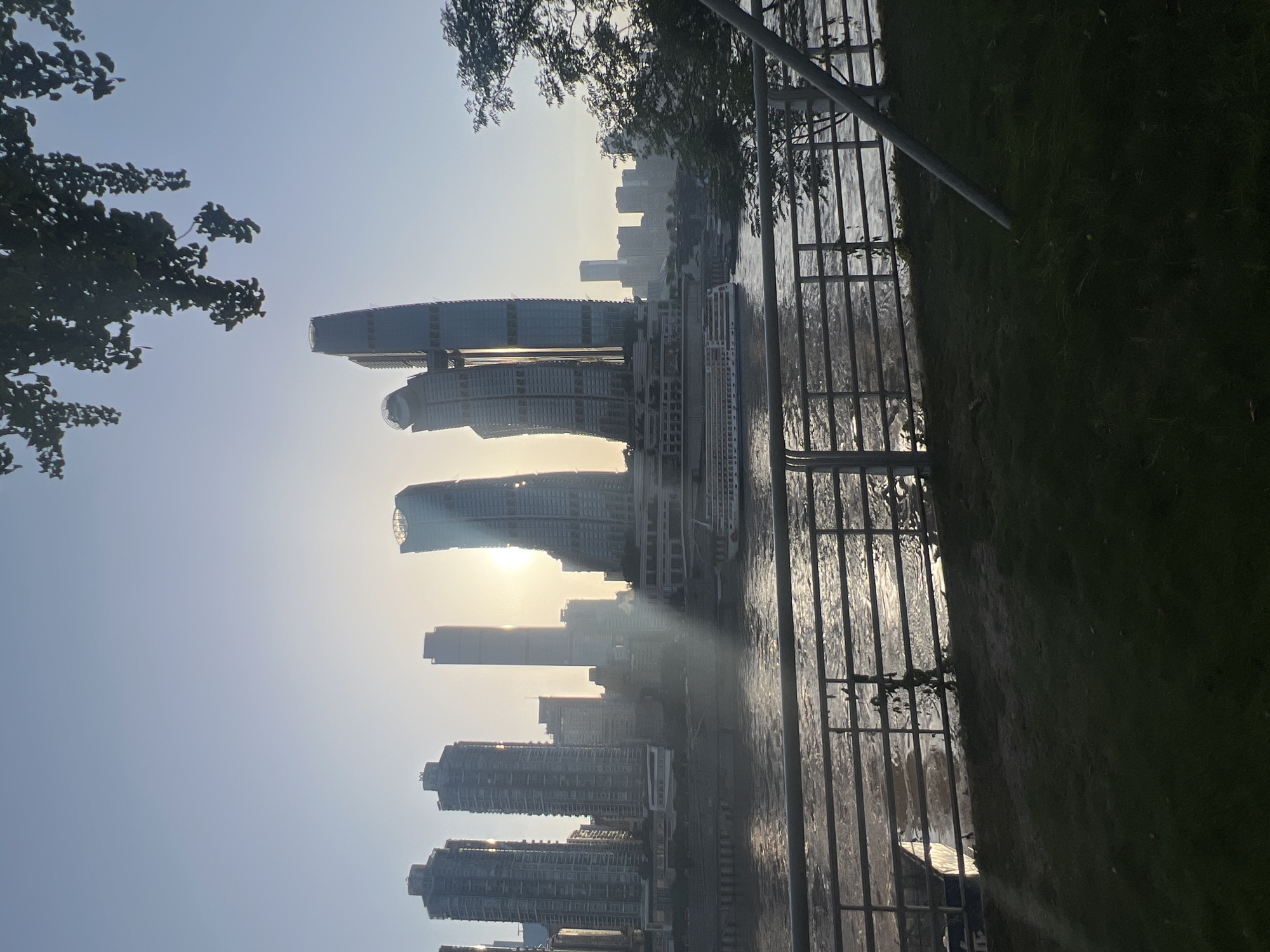 The image depicts a cityscape with a river in the foreground and tall buildings in the background, bathed in the soft light of the setting sun. The location is identified as Nan'an District, China.
In the foreground, a grassy area with a metal railing separates the viewer from the river. The railing is composed of horizontal bars supported by vertical posts. Beyond the railing, a wide river flows, its surface reflecting the sunlight and creating shimmering patterns. A long, white boat with multiple decks is docked or cruising on the river.
The middle ground is dominated by a cluster of modern skyscrapers, some of which have distinctive curved or angular designs. The sun is positioned behind these buildings, casting long rays of light and creating a lens flare effect. The buildings are a mix of glass and concrete structures, with varying heights and architectural styles.
In the background, more buildings of varying sizes and designs can be seen, forming a dense urban skyline. Trees with dark green foliage are visible on the right side of the frame, partially obscuring the buildings and adding a natural element to the urban landscape. The sky is a clear, pale blue, transitioning to a warmer hue near the horizon where the sun is setting. The overall impression is one of a modern city at dusk, with the river and the sunset providing a picturesque backdrop.