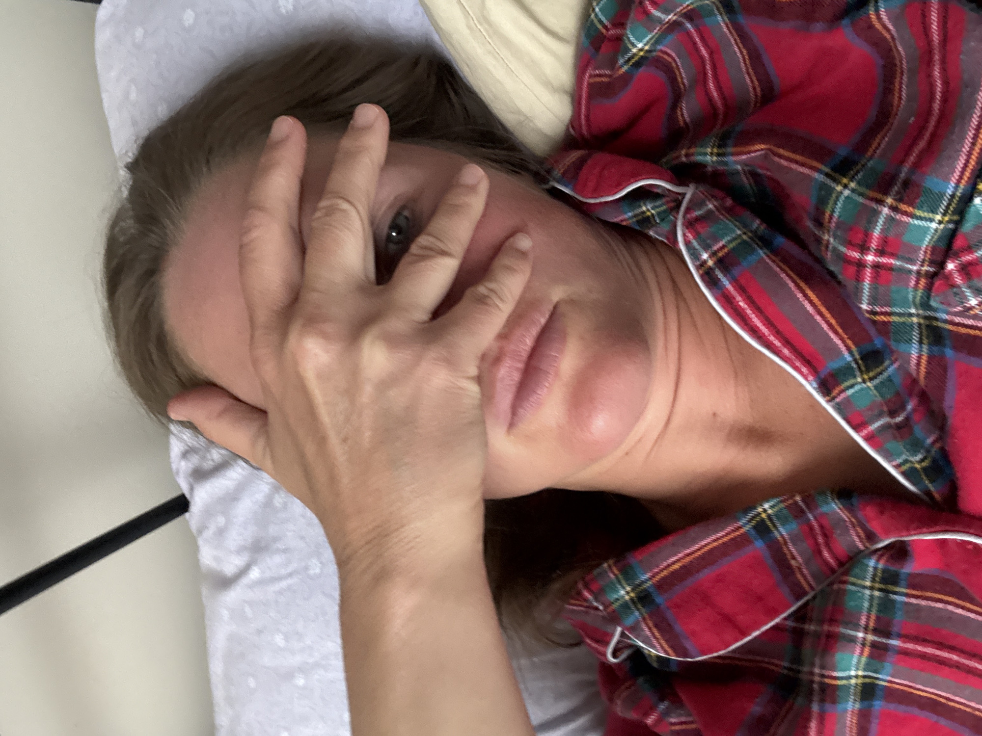 A woman is lying on a light-colored pillow in bed, wearing red plaid pajamas with white piping. Her left hand is covering part of her face. The location cannot be determined to be in Chaikovsky, Russia, or any other specific place from the visual information provided.