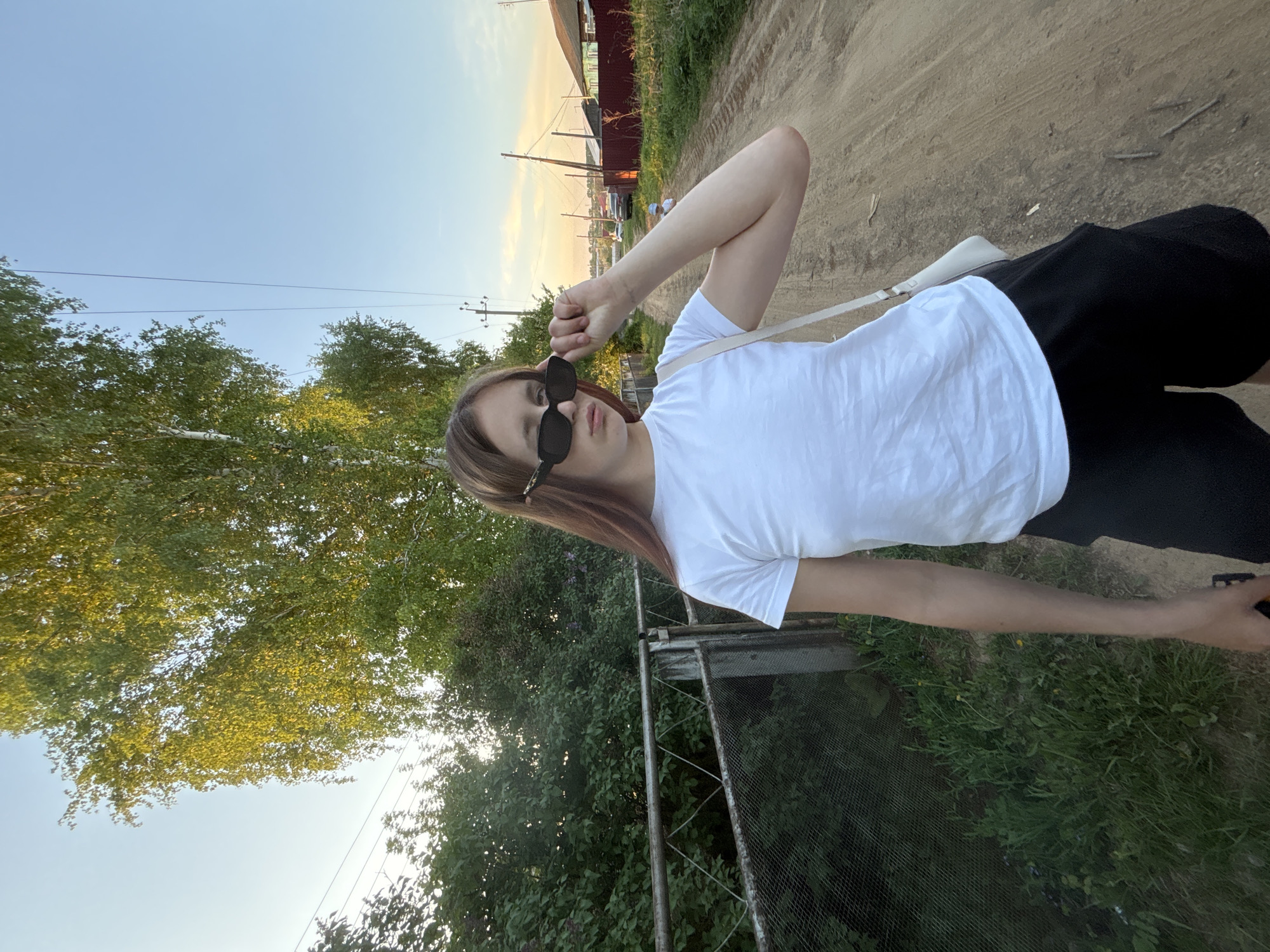 A person stands on a dirt road in Фоки, Russia. They are wearing sunglasses, a white t-shirt, and black pants. A white shoulder bag is slung across their body. A metal chain-link fence runs parallel to the road on the left. Trees are visible behind the fence. A building is visible in the background on the right. The sky is a gradient of blue to orange, suggesting sunset.