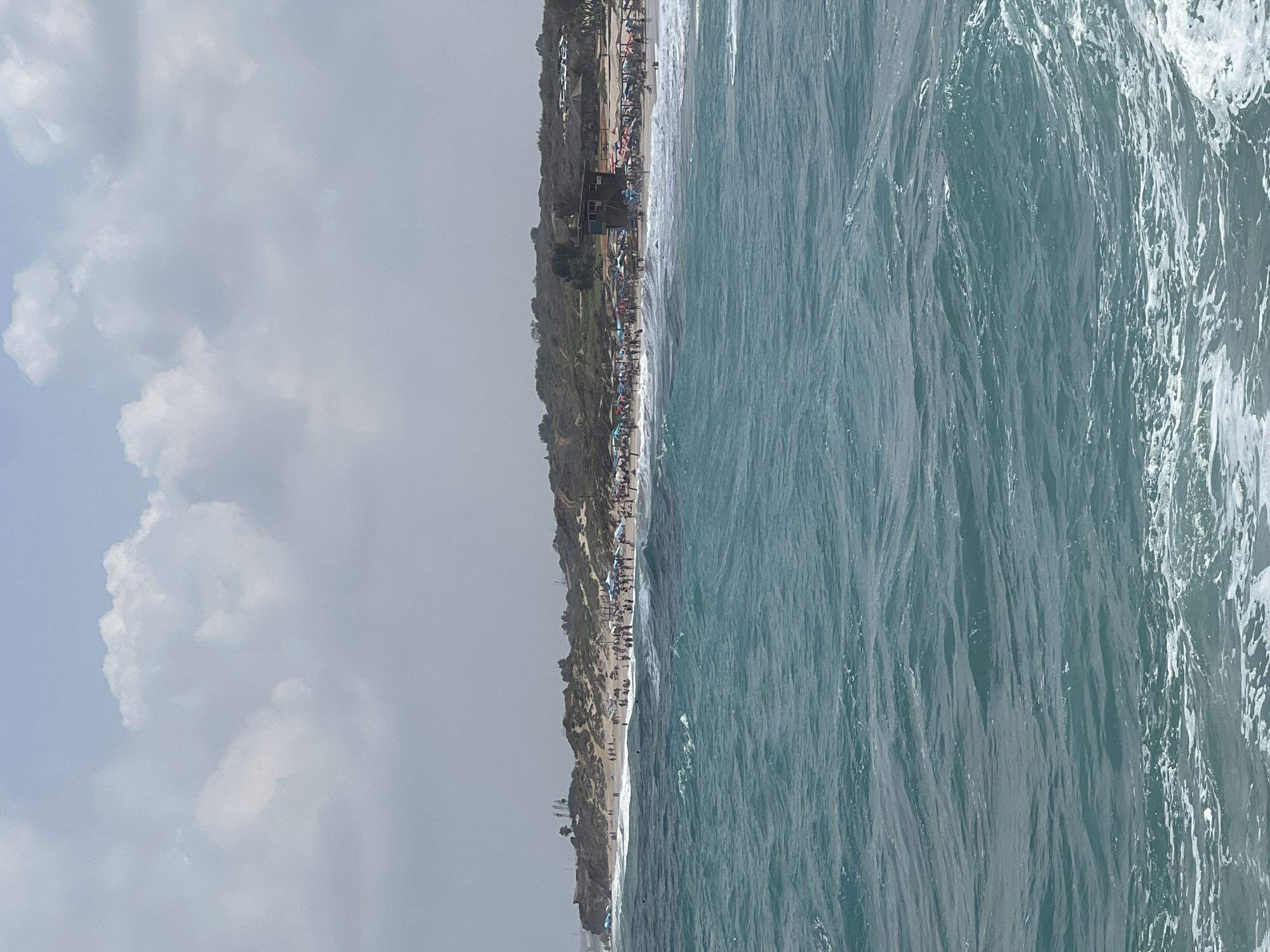 The image depicts a view of the beach and sea at Palmachim, Israel. In the foreground, the turquoise sea is turbulent with white foam and waves. The middle ground shows a crowded beach lined with numerous colorful umbrellas and people sunbathing and walking. Behind the beach, sand dunes covered with vegetation rise up, forming hills. On the dunes, a dark wooden lifeguard station is visible, along with some trees and parked cars. The sky is overcast with light gray and white clouds. The overall scene captures a busy summer day at the beach.